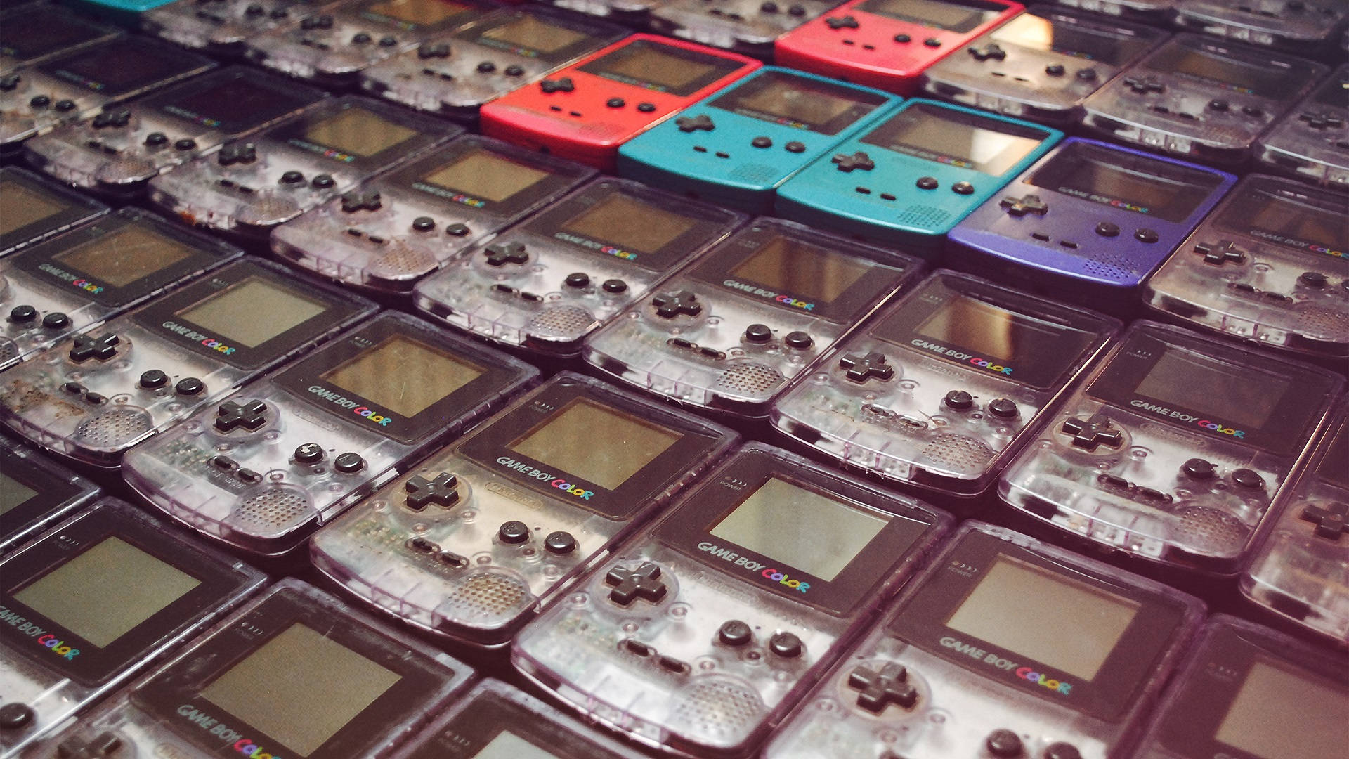 Game Boy Color 各款機型排列 壁紙 — 一個密集的網格，裡面是透明塑膠盒中的 Game Boy Color 掌上遊戲機，紅色、藍色和紫色的主機特別突出。