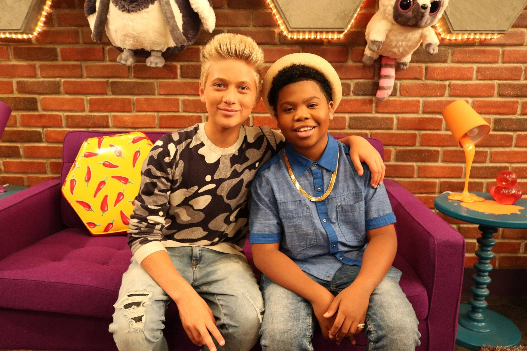 Game Shakers 最佳好友 壁紙 — 兩個微笑的年輕男孩一起坐在紫色沙發上，背景是磚牆，周圍有毛絨玩具和古怪的裝飾。