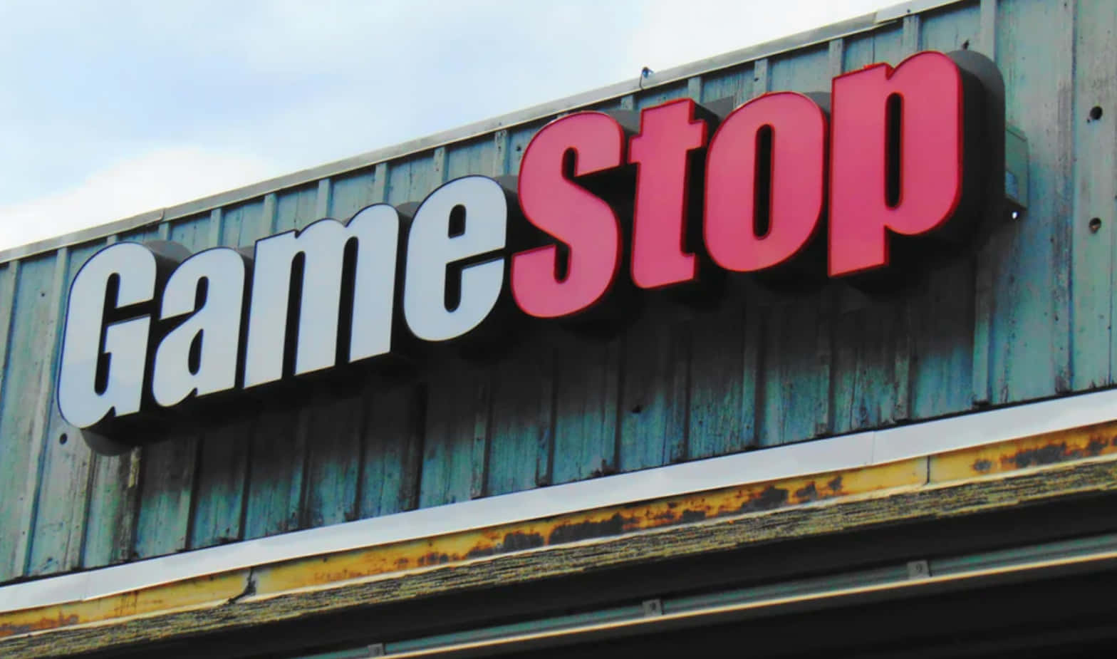 在GameStop購買最新遊戲！ 壁紙 — GameStop 商店招牌，白色和紅色字母安裝在風化的藍色金屬壁板上，天空多雲。
