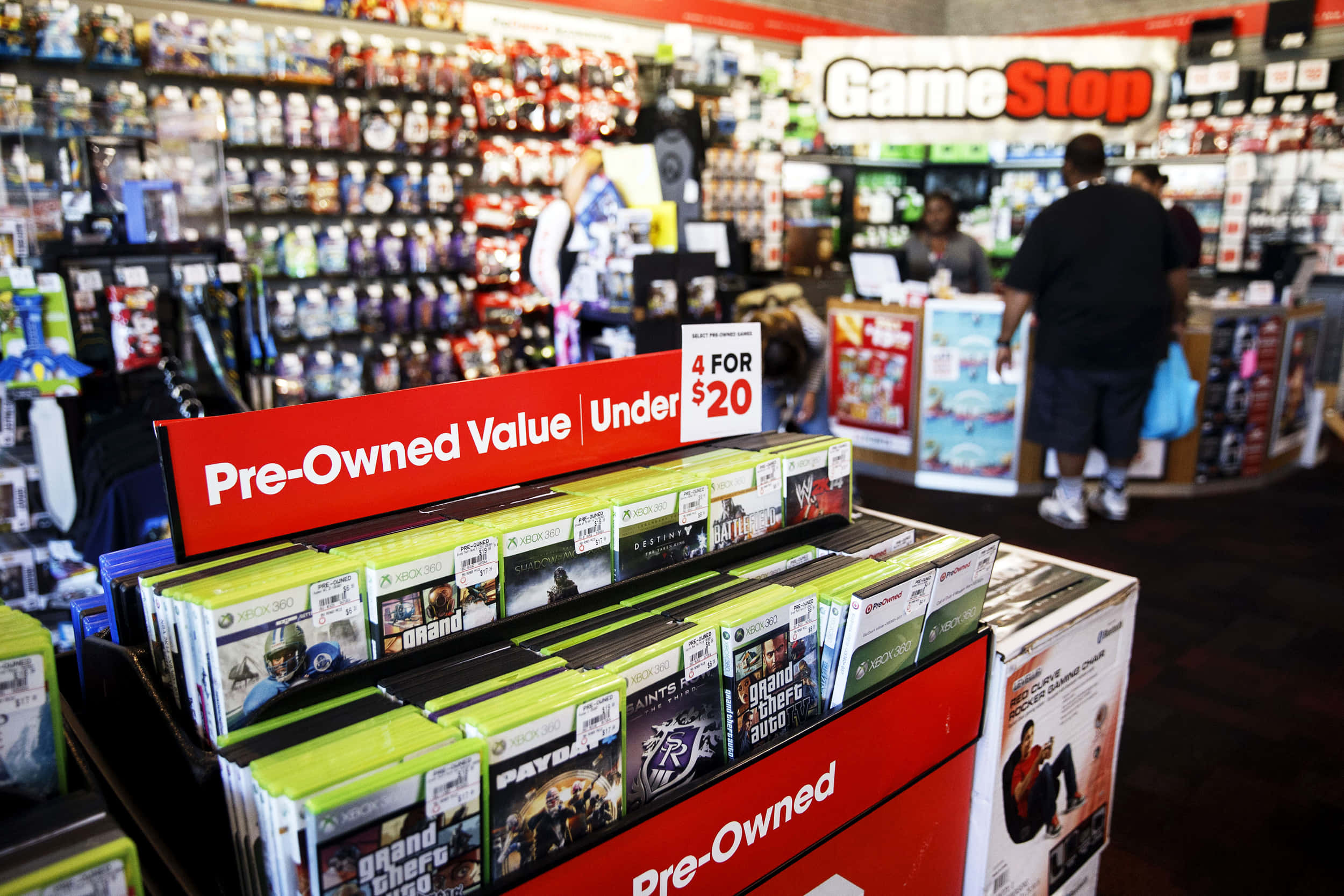 前往 GameStop，準備迎接精彩遊戲時刻 壁紙 — GameStop 商店的貨架上展示著二手 Xbox 遊戲，紅色標誌宣傳四款遊戲低於二十美元。