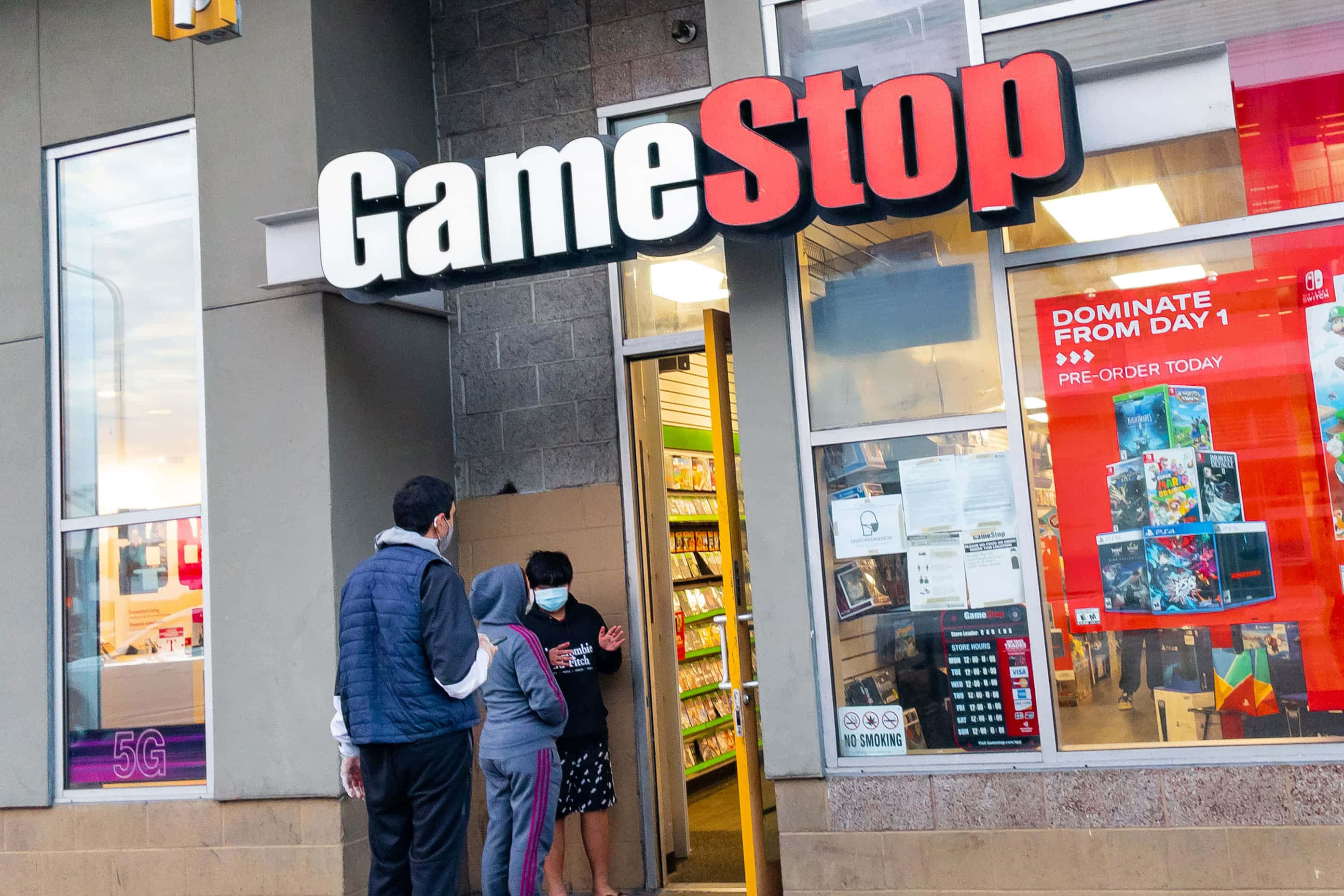 於Gamestop購買最新遊戲 壁紙 — 戴著口罩的人們站在光線明亮的GameStop商店外，窗戶上有紅色標誌和遊戲海報。