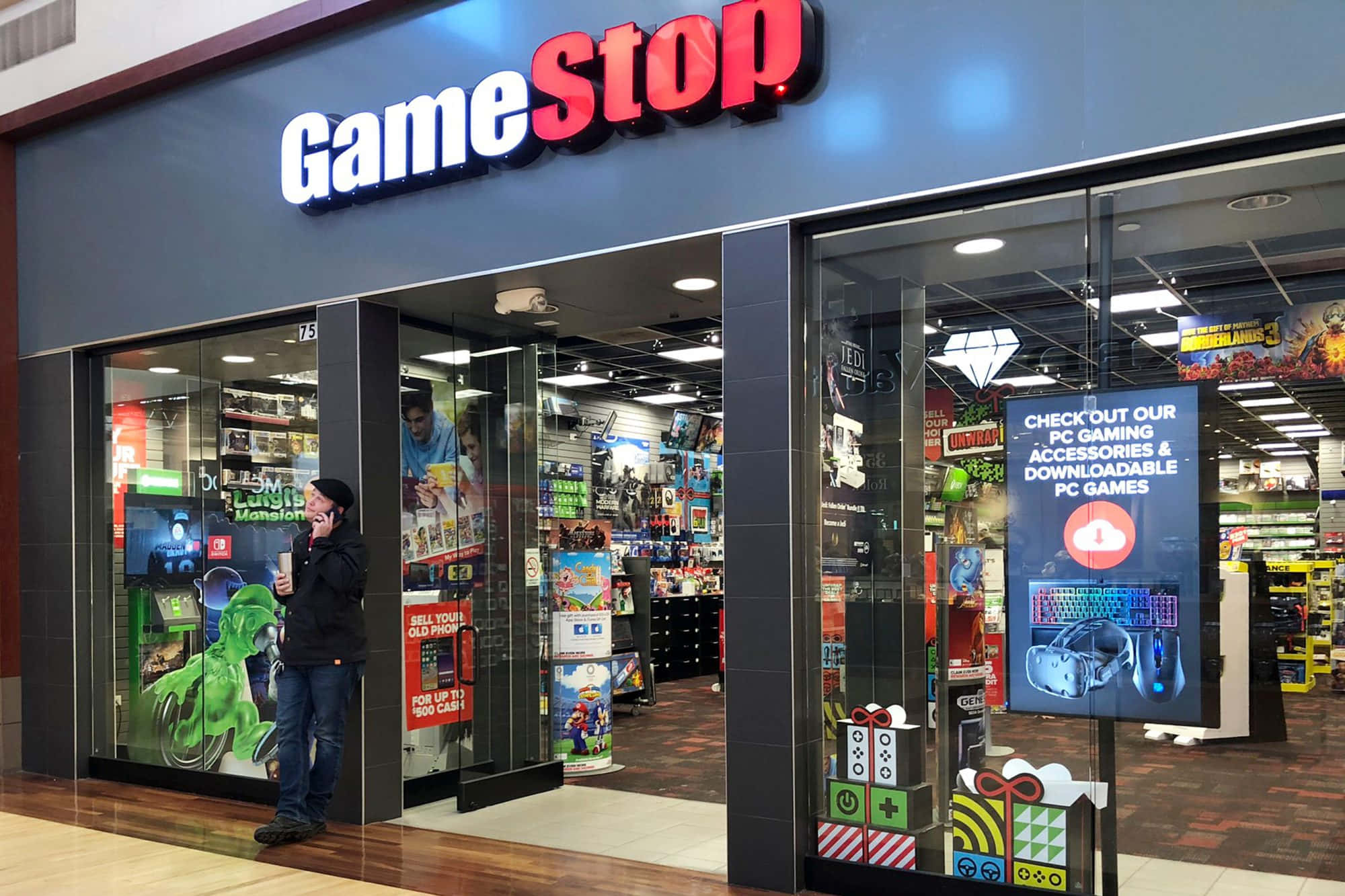 在GameStop購入最新遊戲 壁紙 — 一個人站在燈光明亮的GameStop商店外，玻璃門內可見色彩繽紛的遊戲商品。