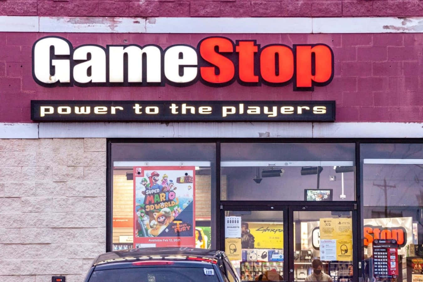 在GameStop選購您最愛的電子遊戲！ 壁紙 — 一間 GameStop 店舖外觀，帶有醒目的紅白招牌，窗戶上可見 Super Mario 3D World 海報。