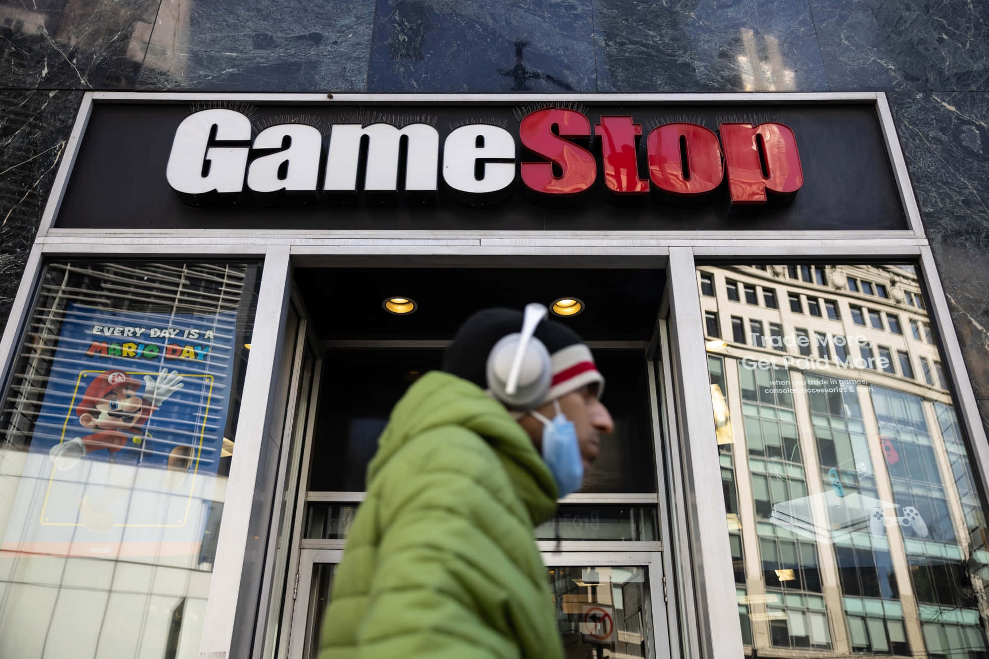 遊戲未來，盡在Gamestop 壁紙 — 一個穿著綠色外套和戴耳機的人走過一間GameStop店，櫥窗裡有瑪利歐海報。
