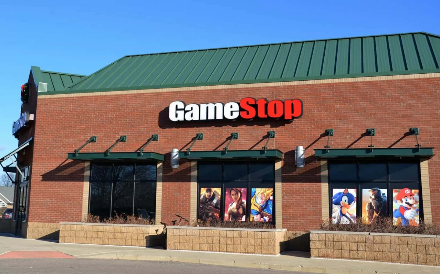 持續遊戲，選擇Gamestop。 壁紙 — 一間明亮的GameStop商店，磚砌外牆、綠色屋頂，窗戶上貼滿色彩繽紛的電子遊戲角色海報，背景是清澈的藍天。