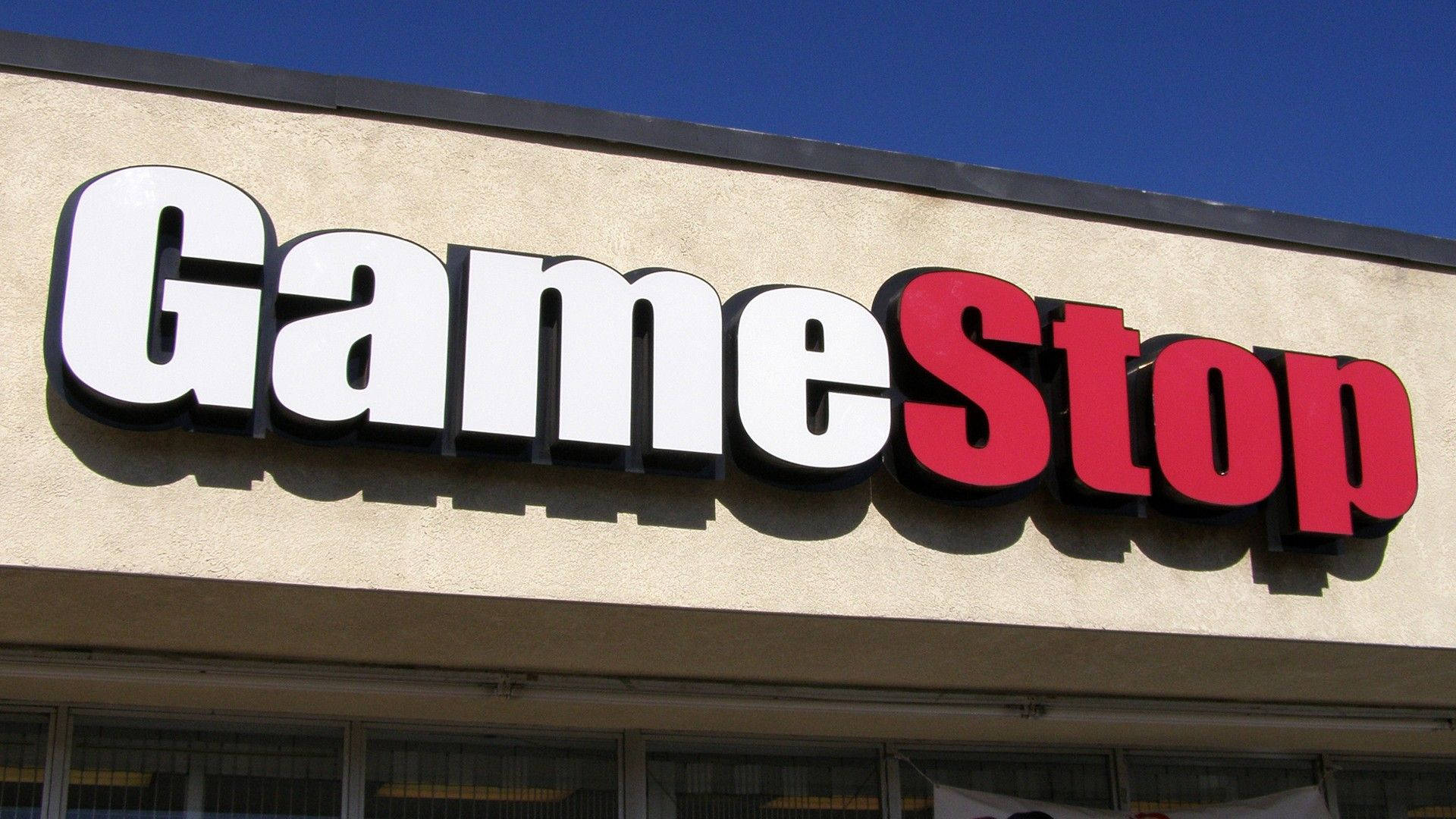 GameStop 標誌招牌 壁紙 — 在晴朗藍天下，米色建築物上安裝了大膽的白色和紅色 GameStop 標誌。