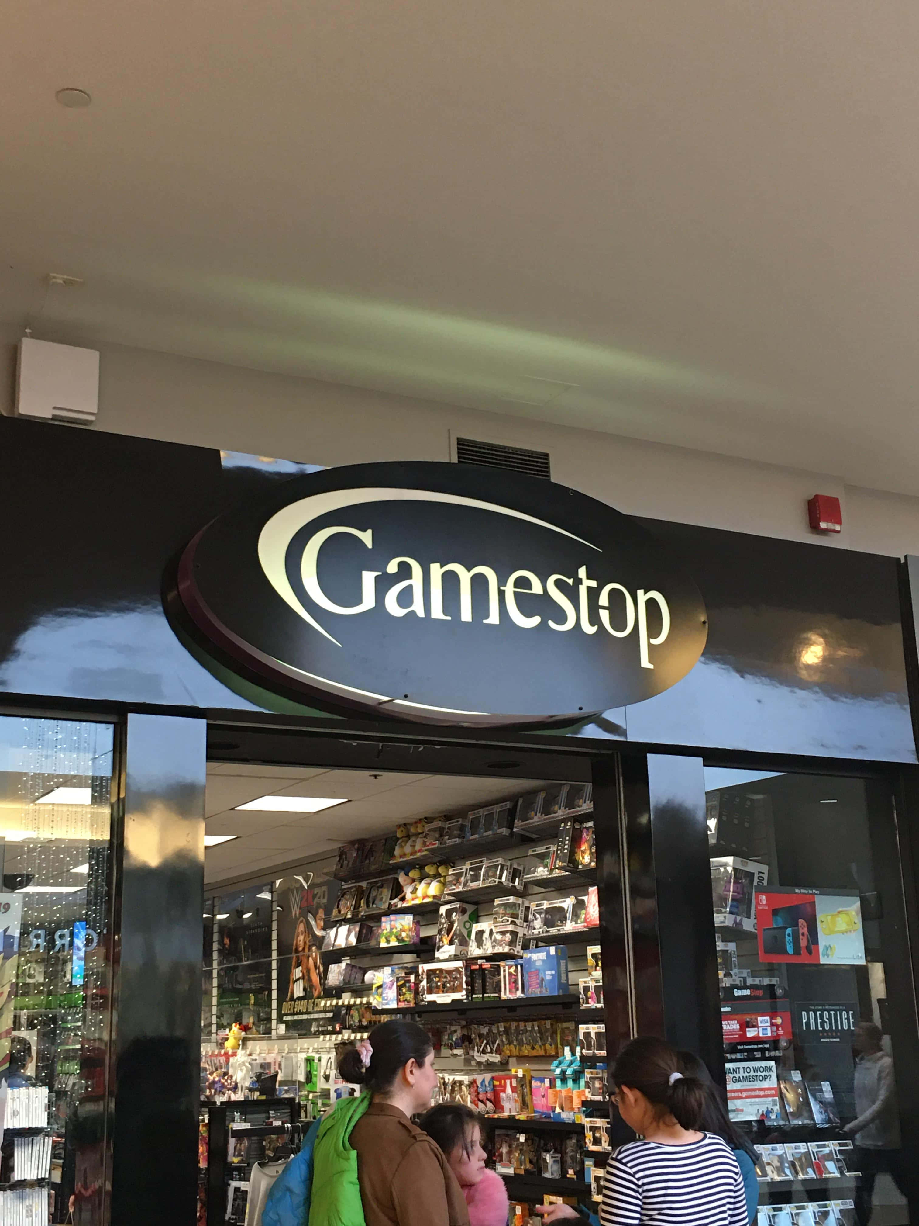 在GameStop購買您最愛的遊戲 壁紙 — GameStop 店舖入口，購物者在明亮的螢光燈下瀏覽電子遊戲和商品。