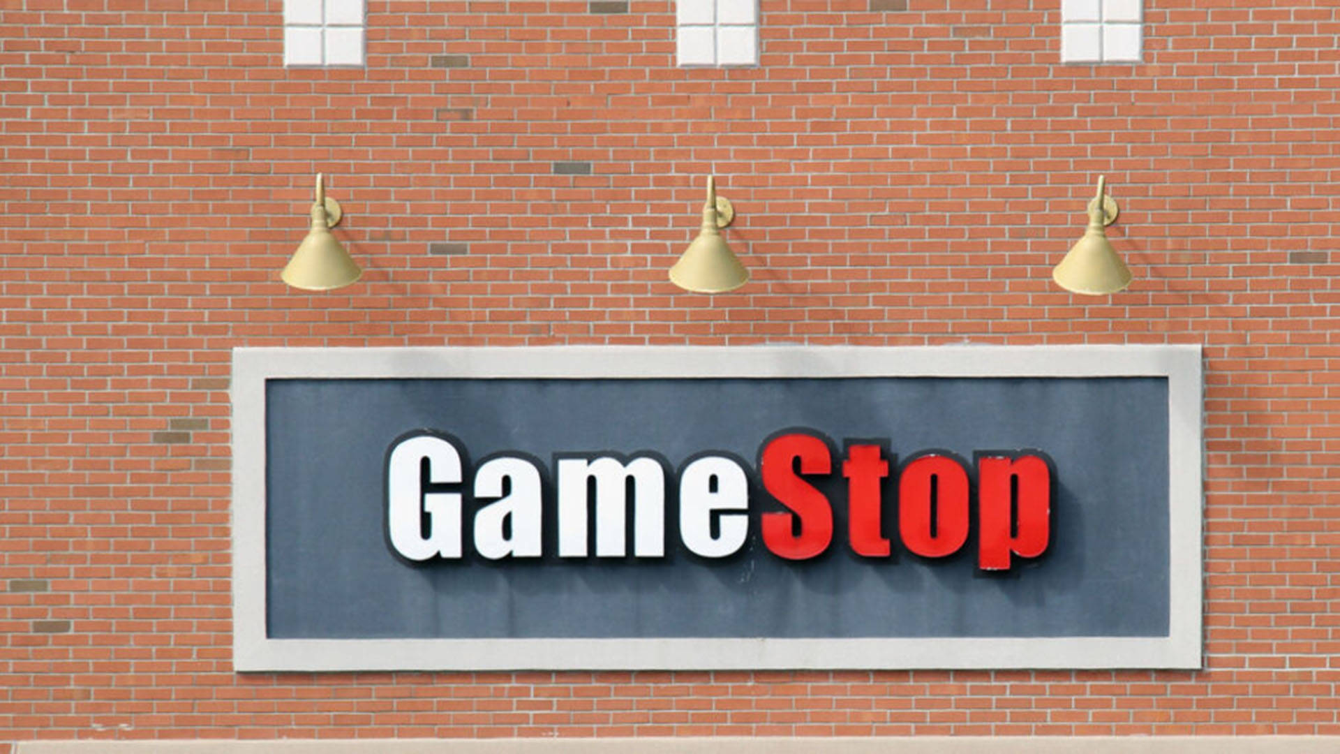 GameStop橙色磚牆招牌 壁紙 — 一個GameStop商店招牌，以粗體白色和紅色字體安裝在紅磚牆上，上方有三盞金色燈光。