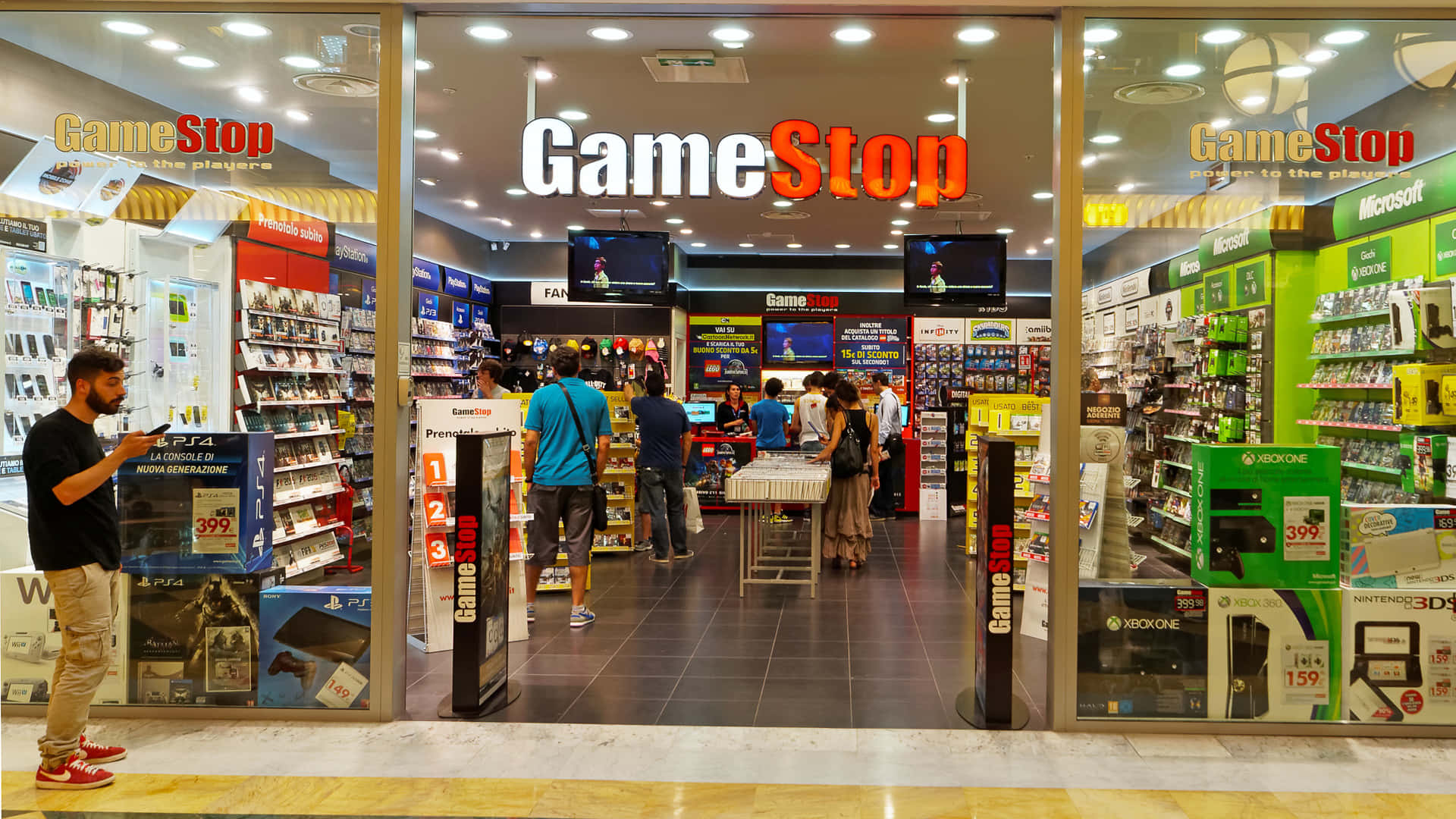 於Gamestop網上搶購最優惠價格 壁紙 — 繁忙的GameStop商店入口，顧客在明亮的頂燈下瀏覽遊戲機和商品。