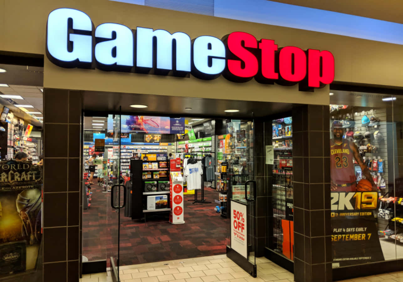 加入GameStop社群，探索最新遊戲動態 壁紙 — 光線充足的GameStop店面，發光招牌和玻璃門展示內部的電子遊戲和商品貨架。