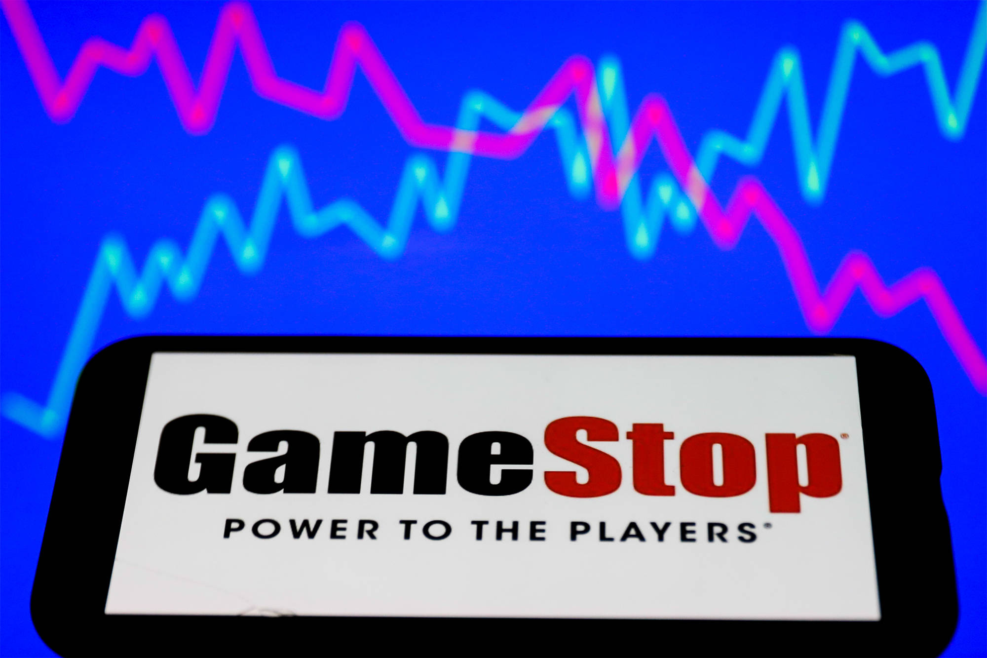 GameStop標誌與藍粉數據圖表 壁紙 — 一部智能手機在充滿活力的藍色背景上顯示GameStop標誌，並帶有發光的粉紅色和青色股票市場圖表。