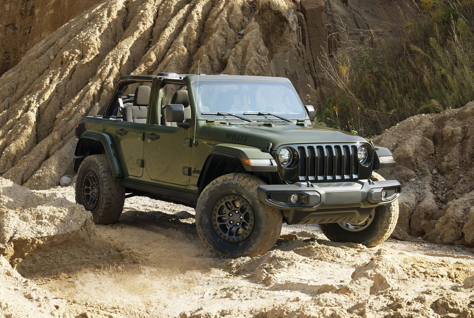 探索你的野性一面 壁紙 — 一輛堅固的橄欖綠色Jeep Wrangler在明亮的日光下，沿著質感岩石 formations 旁邊的陡峭沙坡行駛。