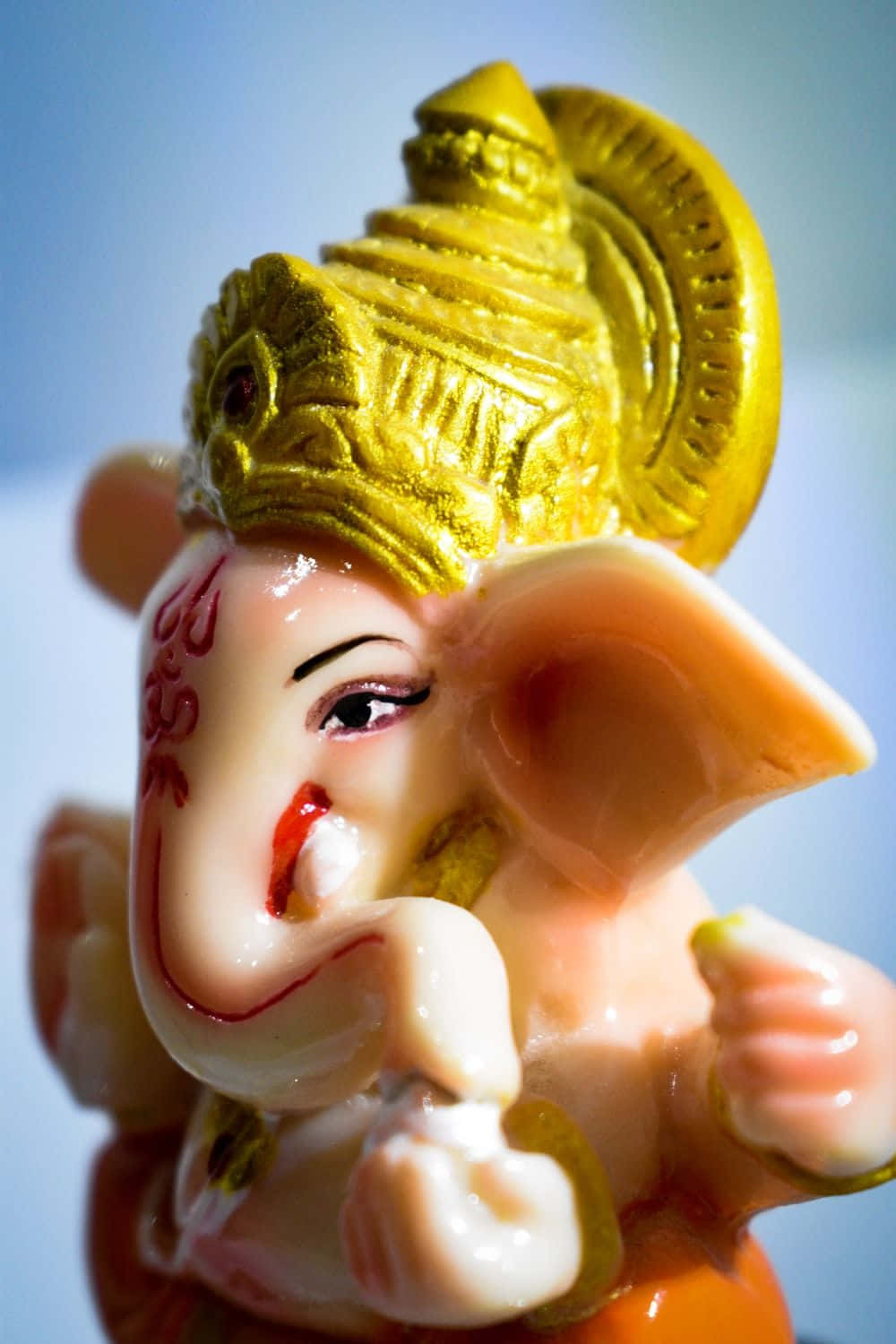 Ganpati Bappa Morya：智慧與愛的象徵 壁紙 — 特寫一個帶有金色皇冠和紅色標記的光澤粉紅色象頭神雕像，背景是柔和的藍色。