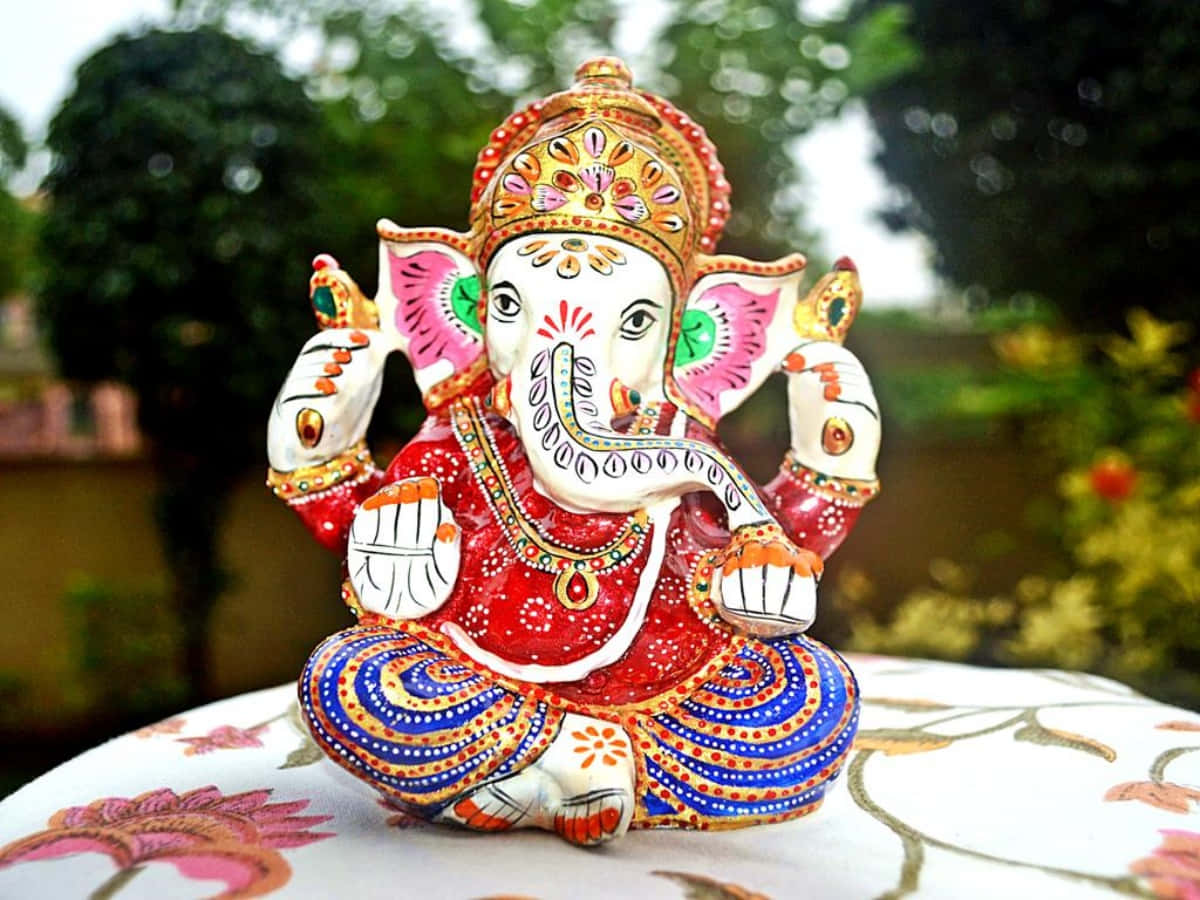 Ganpati Bappa 印度迷你神像圖像 壁紙 — 一尊色彩鮮豔的象神雕像安詳地坐在花卉桌布上，背景是柔和模糊的花園。