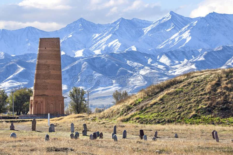 Kyrgyzstan 桌布 壁紙 — 一座高磚塔矗立在草地上，散落著墓碑，天空多雲，背景是白雪覆蓋的山脈。
