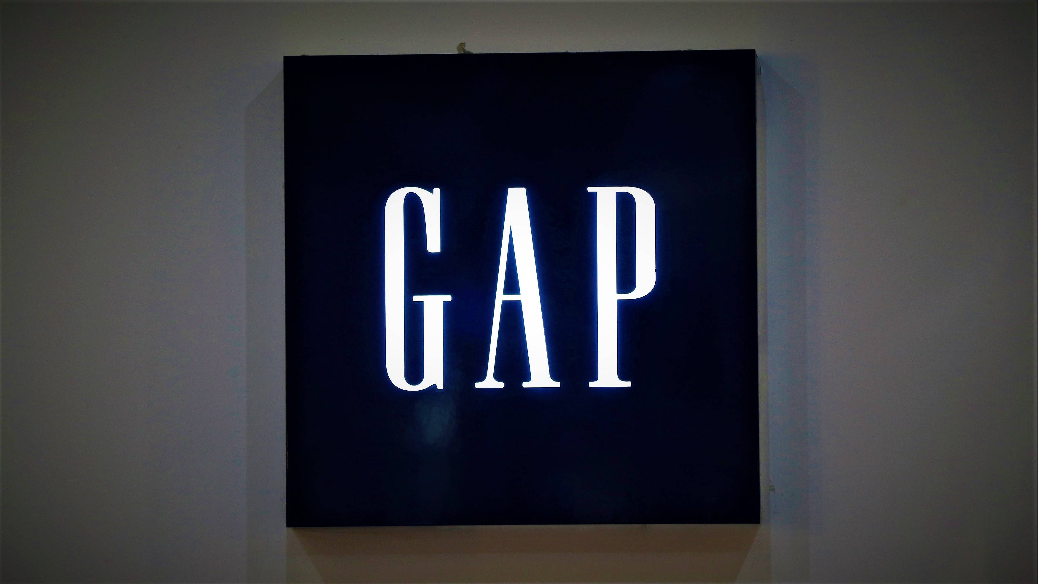 Gap 標誌牆上招牌 壁紙 — 一個發光的白色GAP標誌招牌，安裝在昏暗室內空間的素牆上。