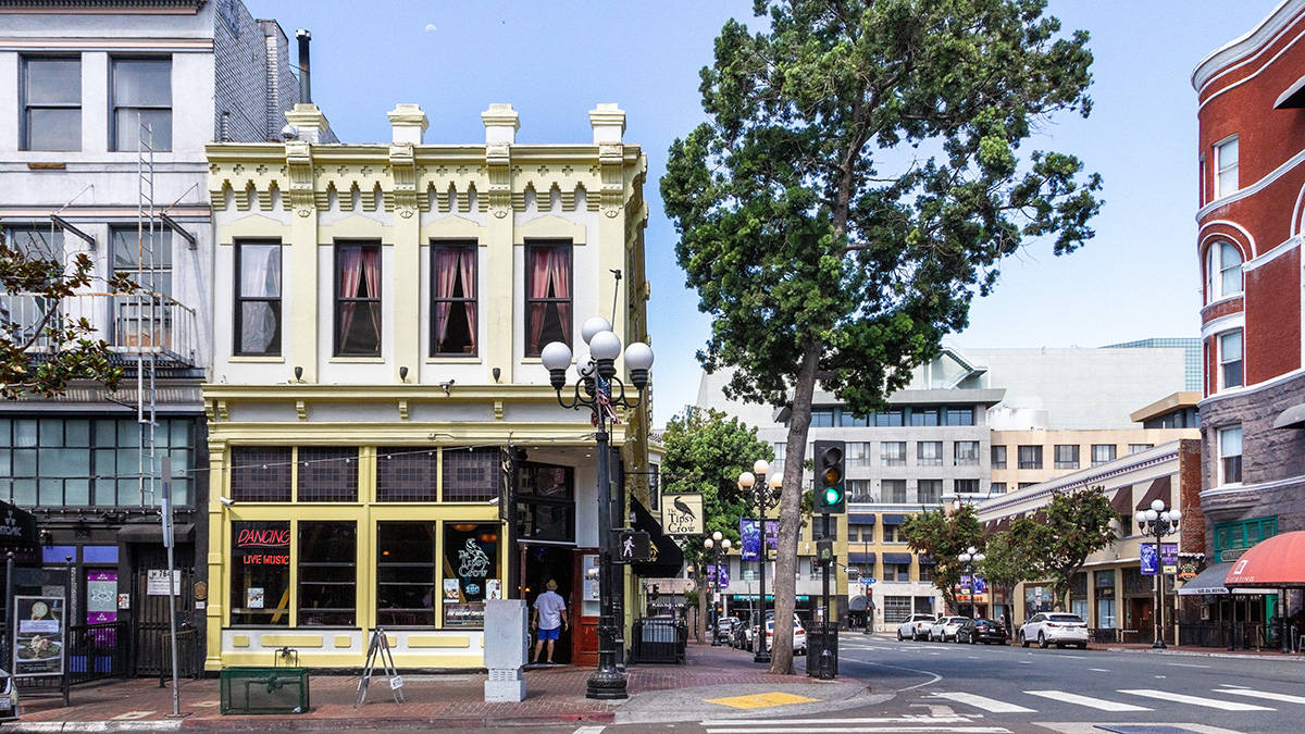 聖地牙哥歷史_gaslamp區風光 壁紙 — 一個陽光明媚的街角，有一座歷史悠久的黃色建築，配有紅色窗簾和跳舞標誌，周圍環繞著茂密的綠樹和經典的路燈。