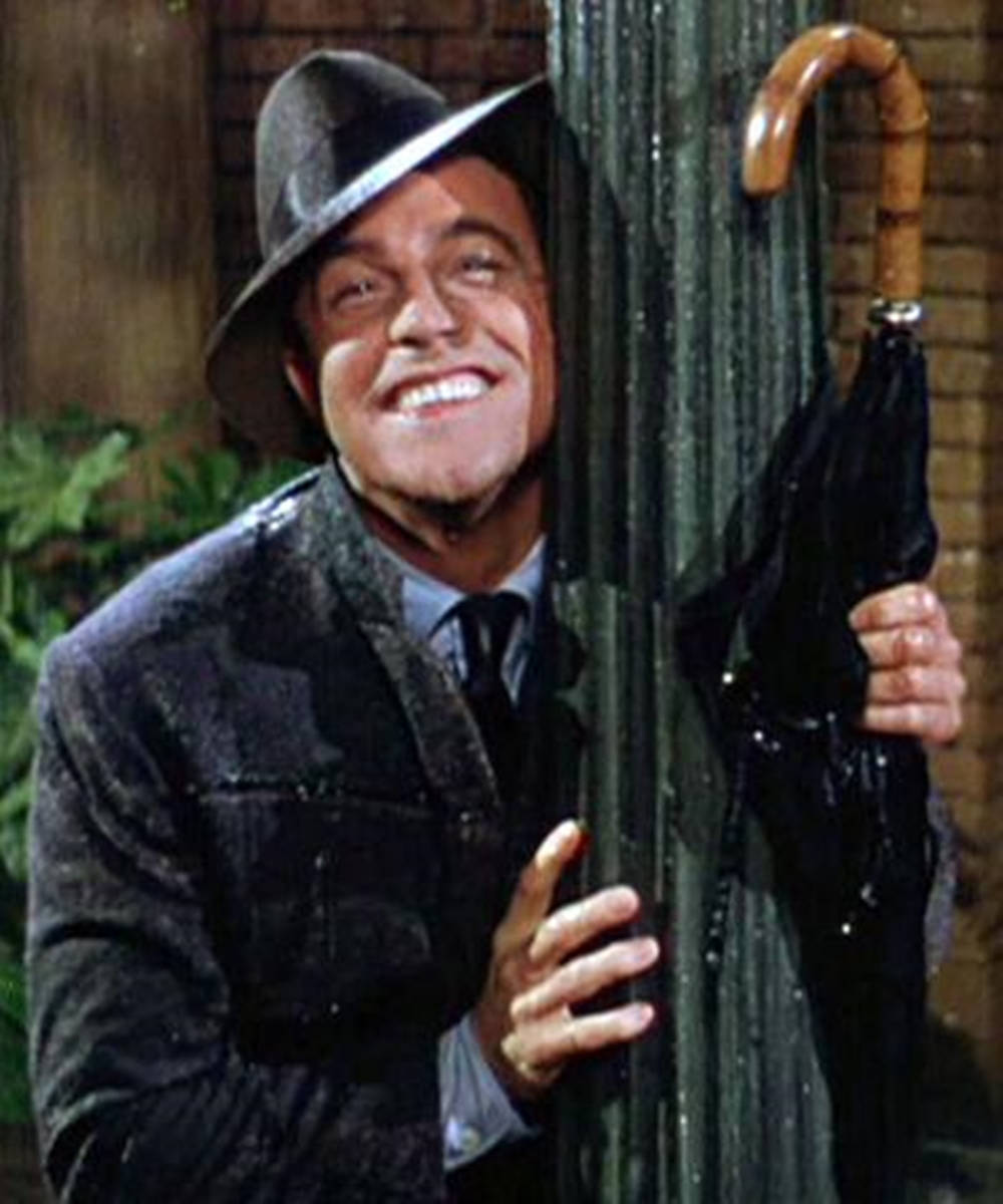 Gene Kelly 拥抱欄杆 壁紙 — 一個戴著紳士帽和雨衣的開朗男子，在濕滑的柱子後面拿著濕雨傘微笑。