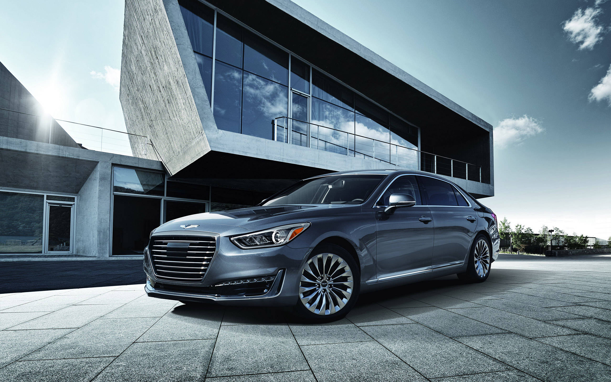 高畫質藍鋼色Genesis G90豪華轎車 壁紙 — 一輛時尚的銀色轎車停泊在瓷磚廣場上，前方是現代玻璃與混凝土建築，天空晴朗。