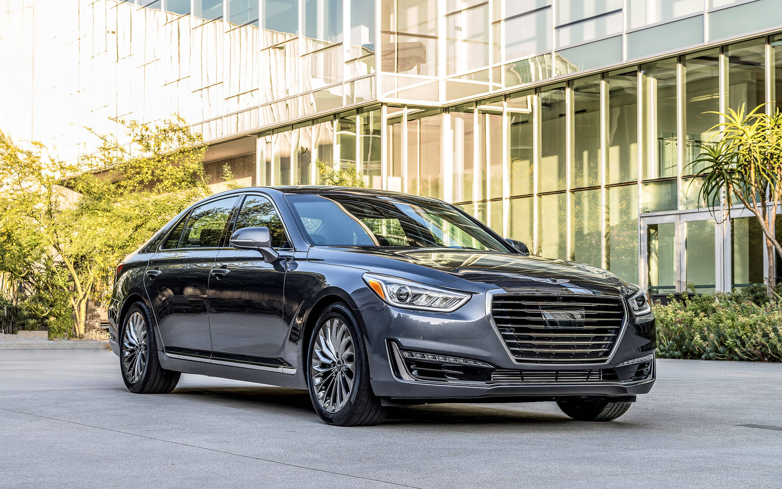 鋼灰色 Genesis G90 壁紙 — 一架時尚的深灰色豪華轎車停泊在現代玻璃建築前，陽光在其拋光表面上投射出反射。
