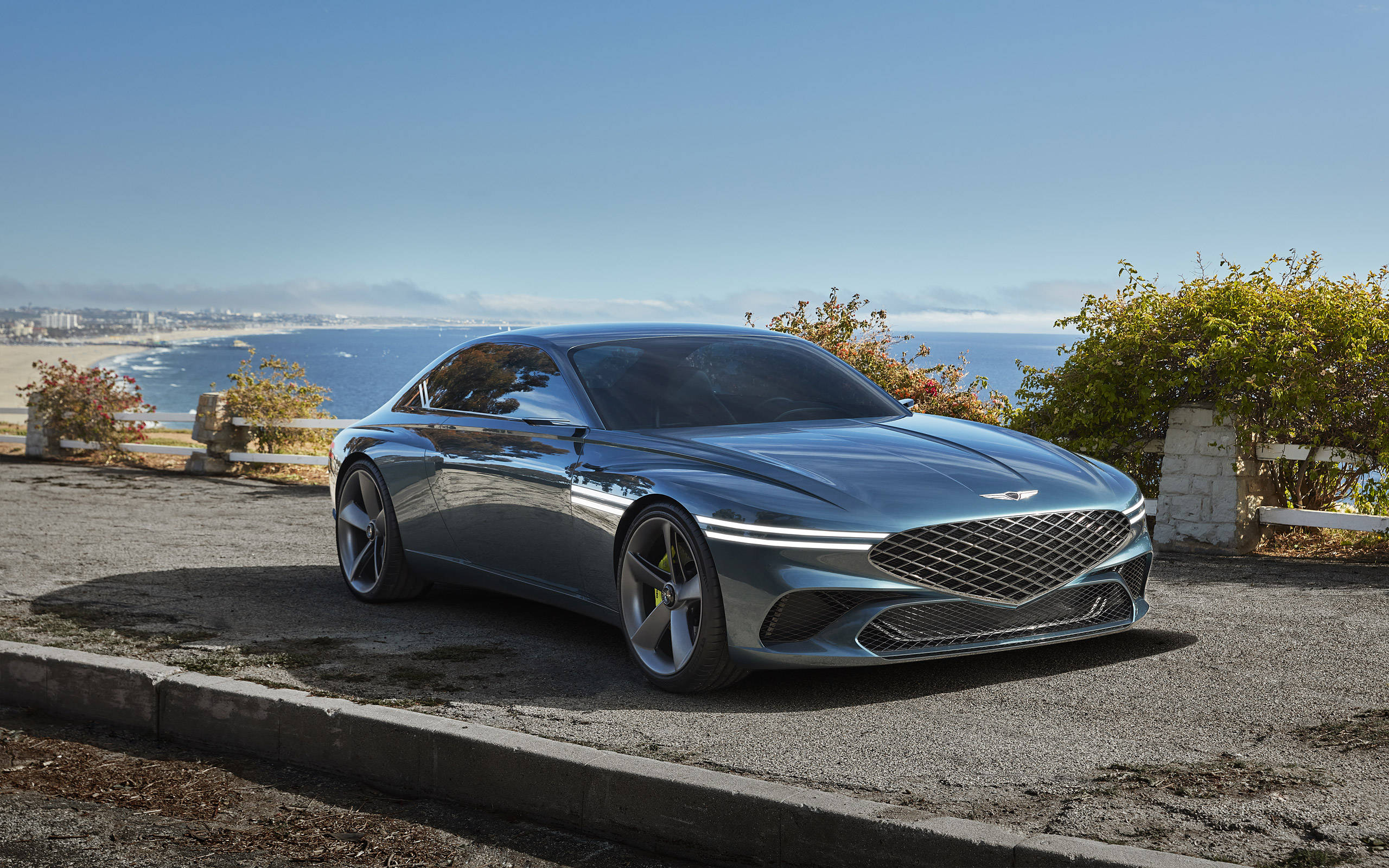 「Genesis X - 讓奢華引領方向」 壁紙 — 一輛時尚的銀色跑車在明亮的陽光下閃閃發光，位於海岸懸崖上，俯瞰藍色海洋和遠處的海灘小鎮。