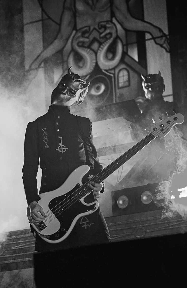 Ghost Band黑白吉他手照片 壁紙 — 一位戴著惡魔面具的音樂家在煙霧彌漫的舞台上彈奏低音結他，背景是惡魔彩繪玻璃。