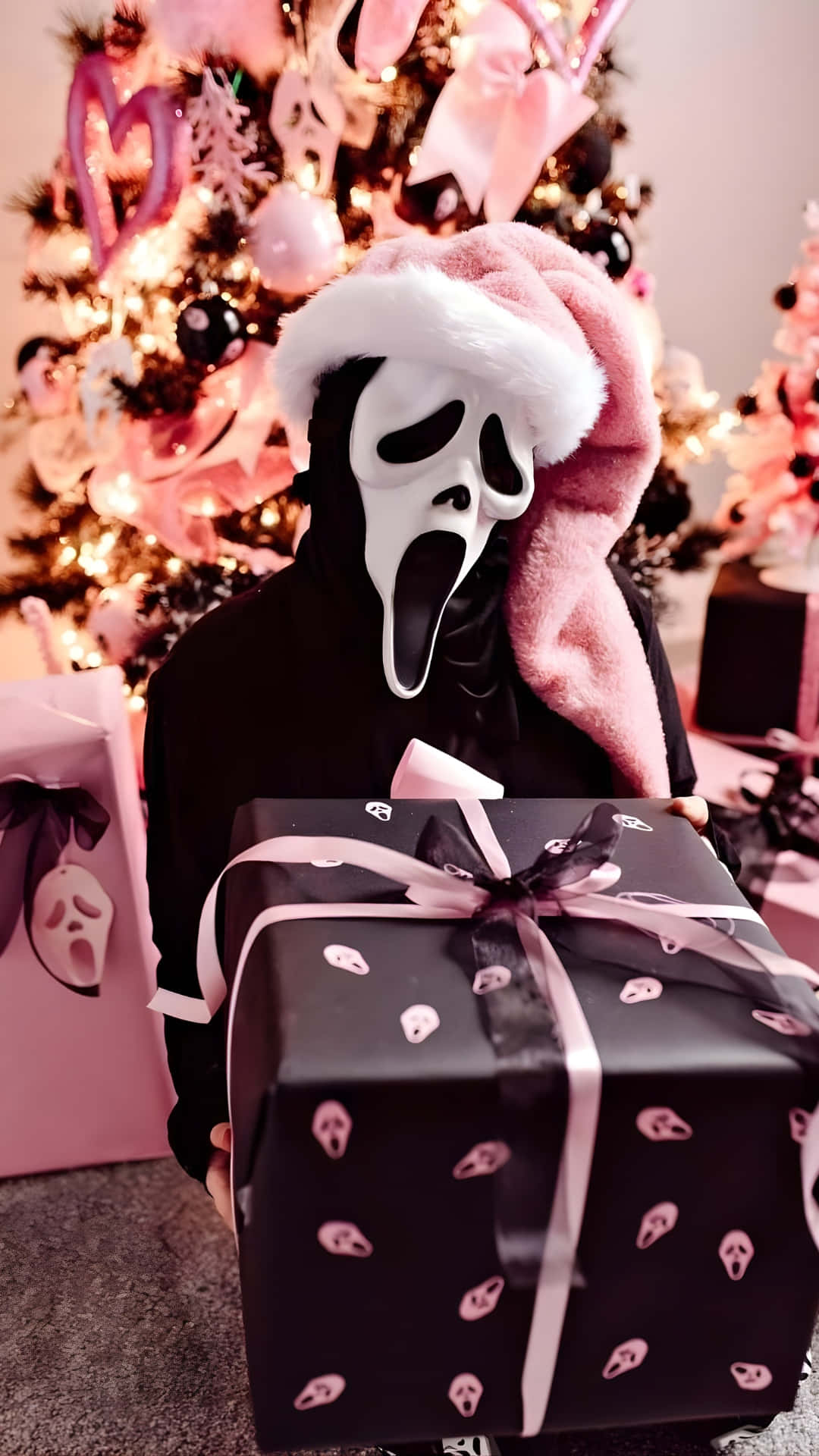  壁紙 — Ghostface 戴住粉紅色聖誕帽，喺粉紅色同黑色裝飾嘅聖誕樹前面，拎住一個有粉紅色骷髏圖案嘅黑色禮物盒。