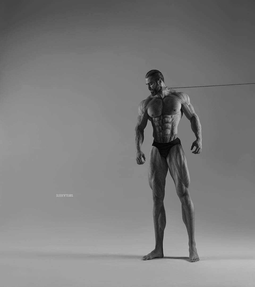 Giga Chad 肌肉模特兒自信擺姿 壁紙 — 一個肌肉發達的男人赤腳站在簡約的工作室中，他清晰的體格在戲劇性的燈光下，以純灰色背景襯托出來。