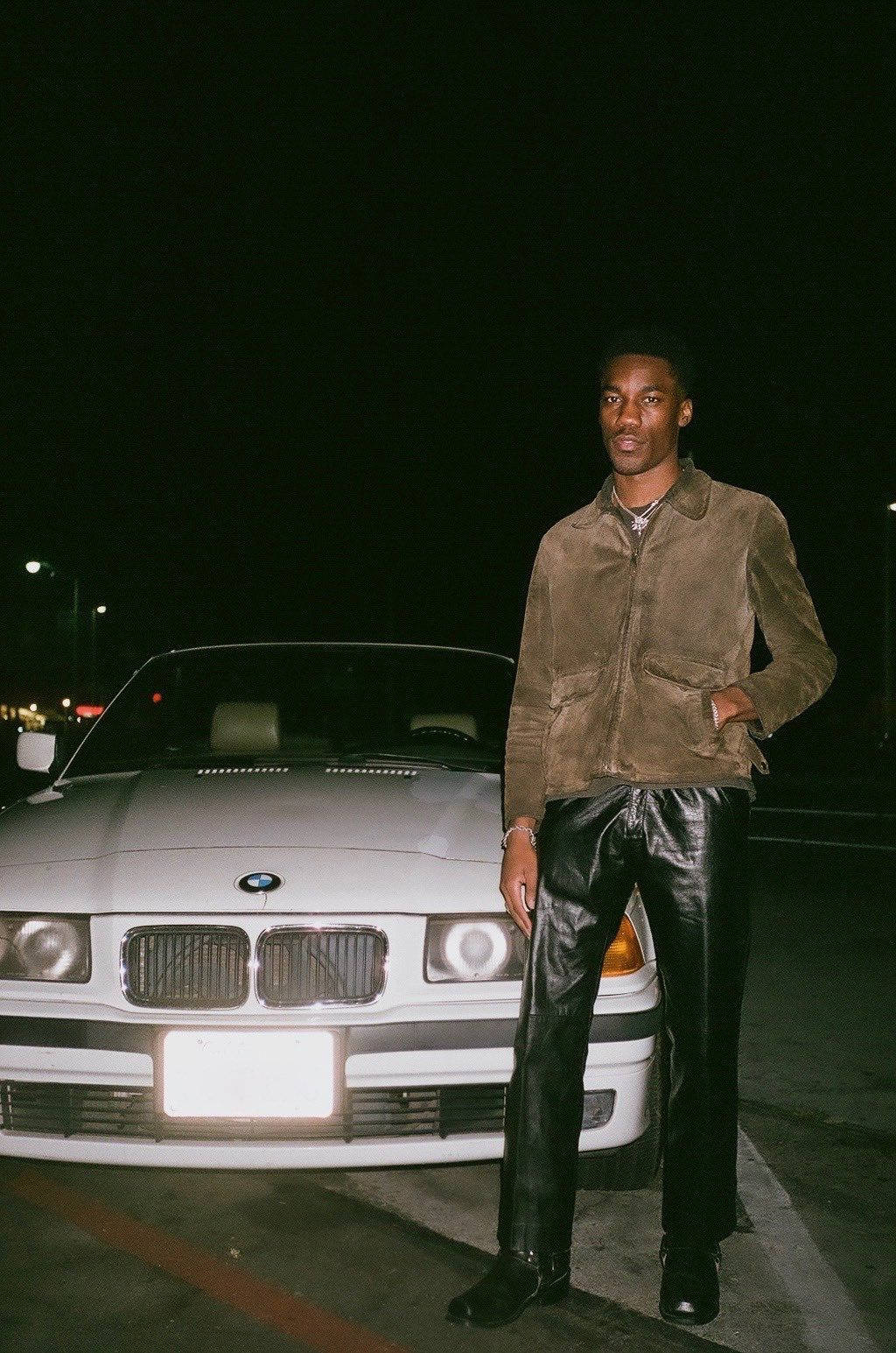 Giveon 與 BMW 車款 壁紙 — 一名身穿棕色麂皮外套和黑色皮褲的男子，在夜晚的路燈下自信地站在一輛白色寶馬旁。