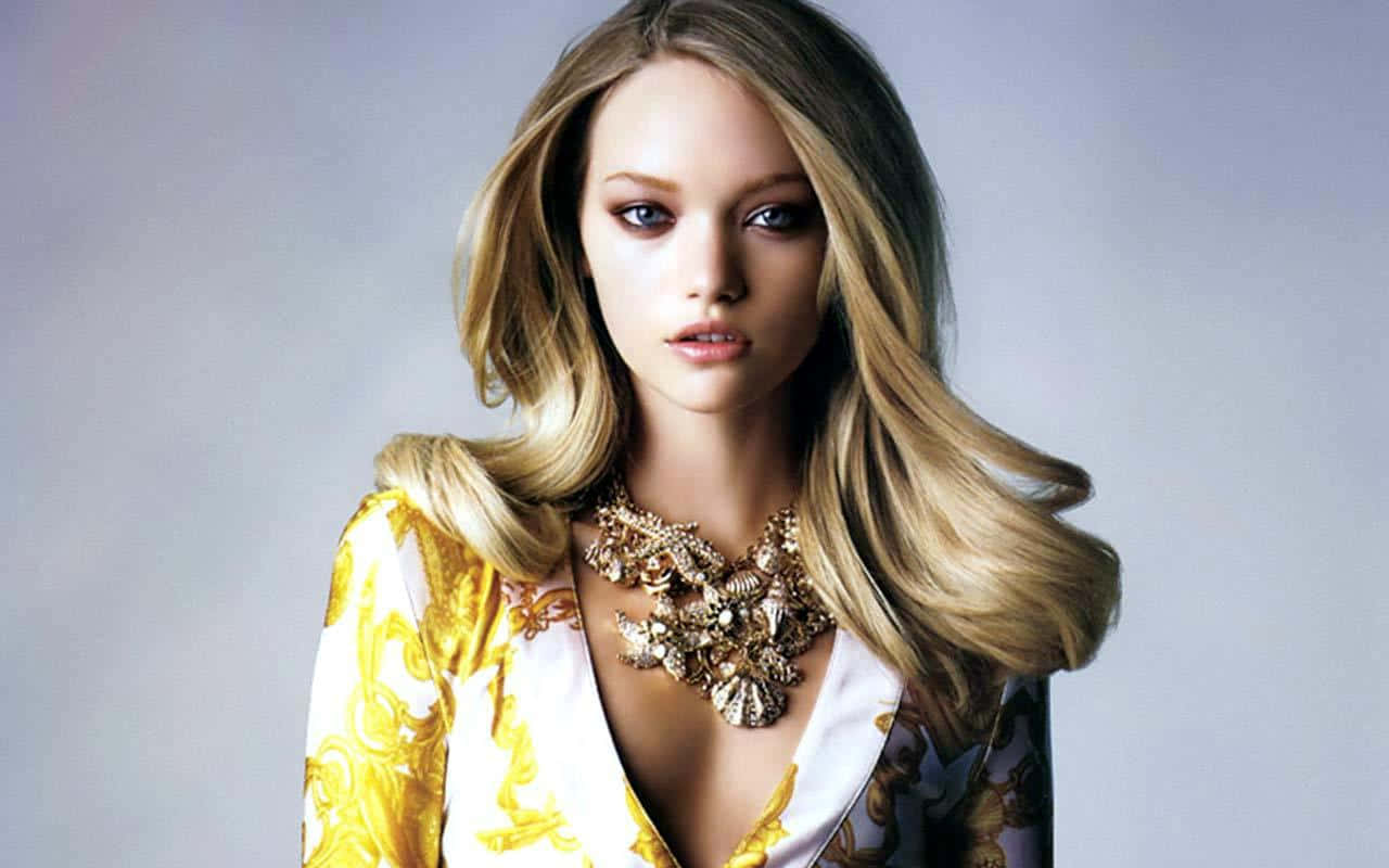 Gemma Ward 桌布 壁紙 — 一位迷人的金髮模特兒，擁有引人注目的藍色眼睛，穿著金色和白色圖案的上衣和華麗的金色項鍊，背景是柔和的灰色攝影棚。