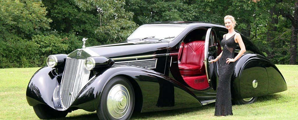 Rolls Royce Phantom Drophead Coupe 桌布 壁紙 — 一位身穿黑色連衣裙的魅力女性站在草地上，旁邊是一輛線條流暢的黑色復古車，車內有紅色皮革內飾。