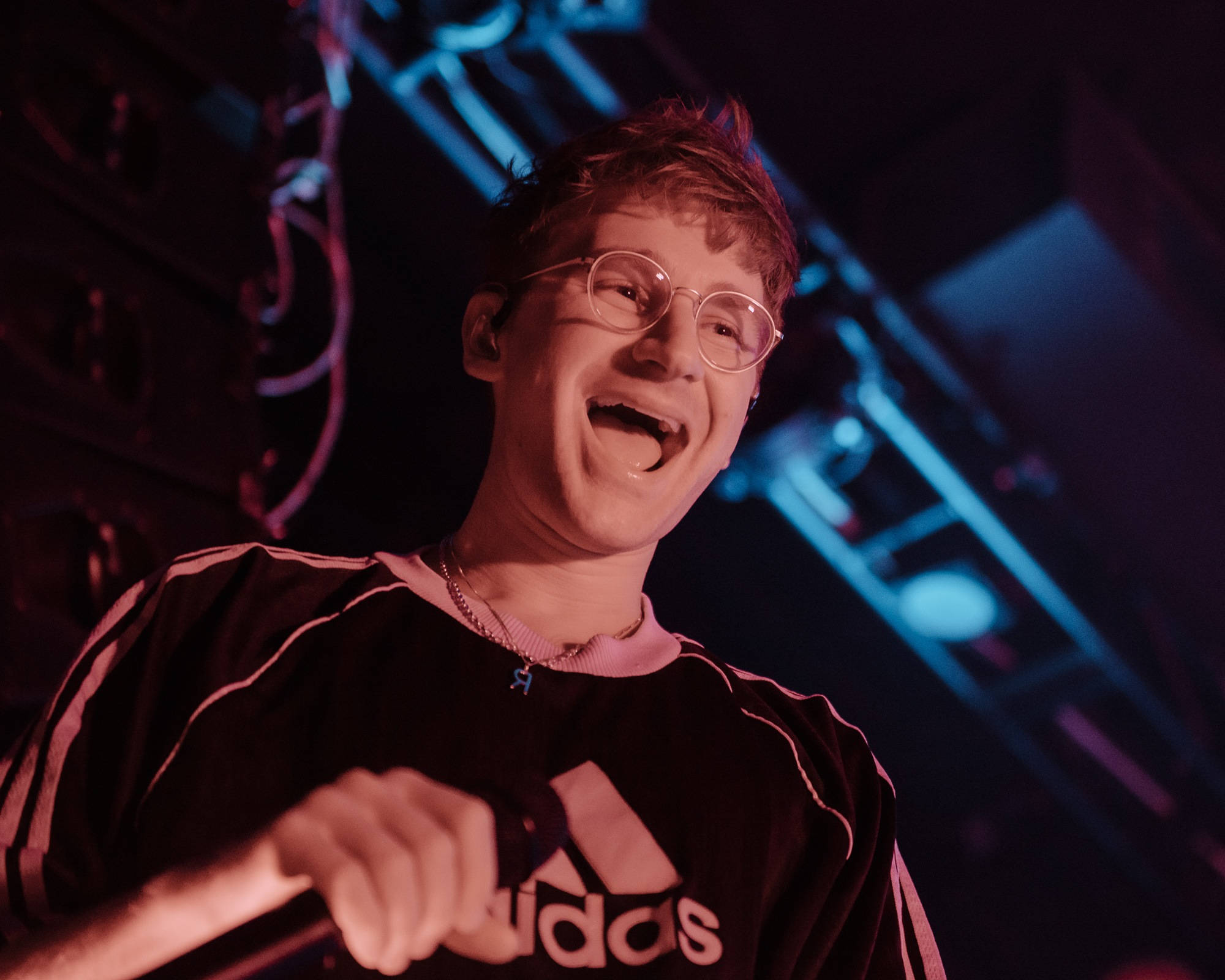 Glass Animals Dave Bayley 微笑瞬間 壁紙 — 一個戴眼鏡的年輕人在昏暗的場地中，在鮮豔的紅色和藍色舞台燈光下開懷大笑。