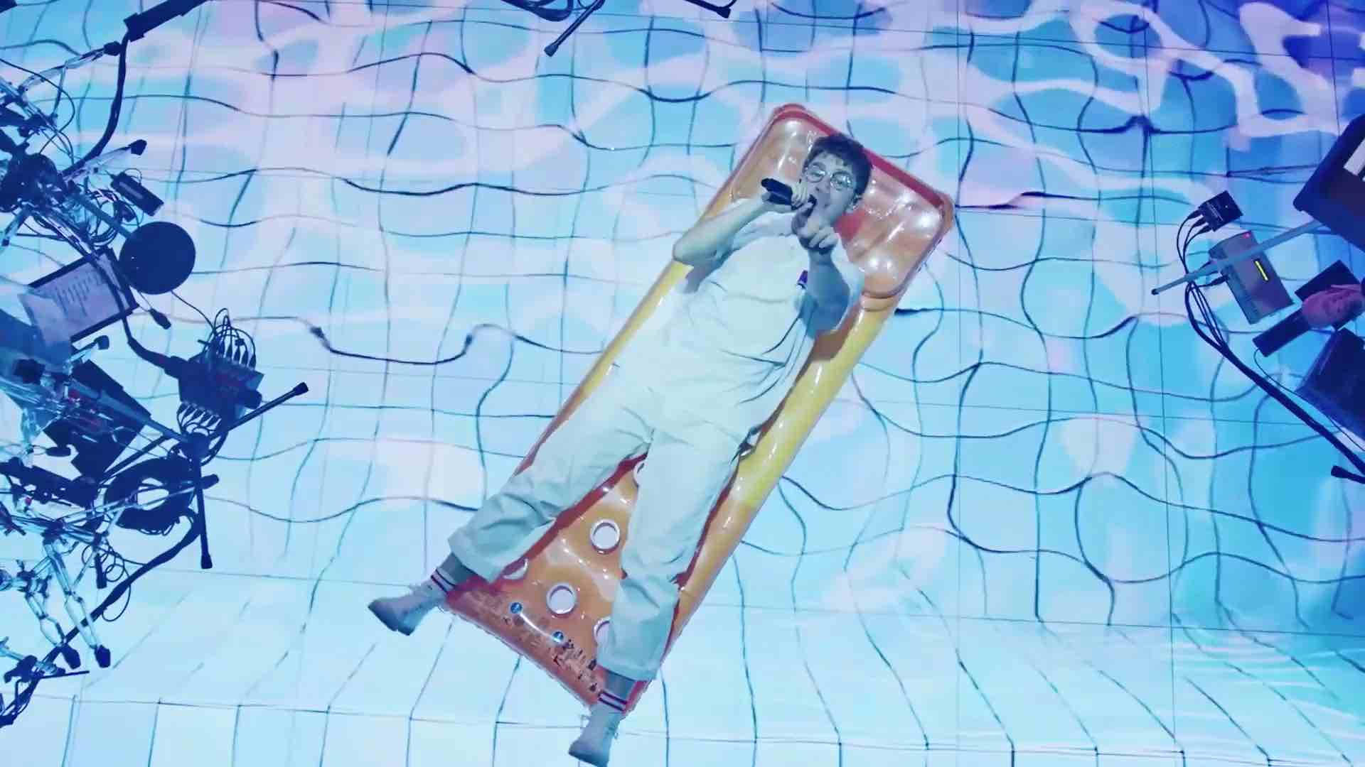 Glass Animals Dave Bayley 演唱《IASIL》 壁紙 — 一名男子躺在橙色充氣筏上，漂浮在藍色瓷磚泳池地板上，周圍環繞著音樂設備。