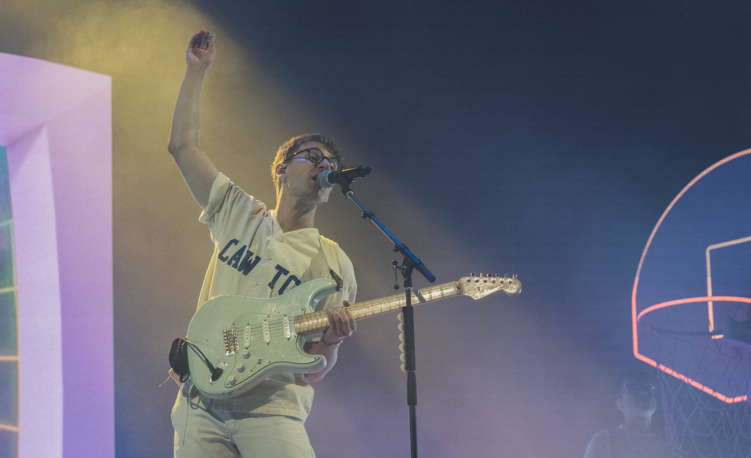 Glass Animals Dave Bayley 舞台彈電吉他 壁紙 — 一位身穿白色服裝的音樂家熱情地彈奏淺綠色電結他，並在舞台燈光下對著咪高峰唱歌，背景有一個發光的籃球框。