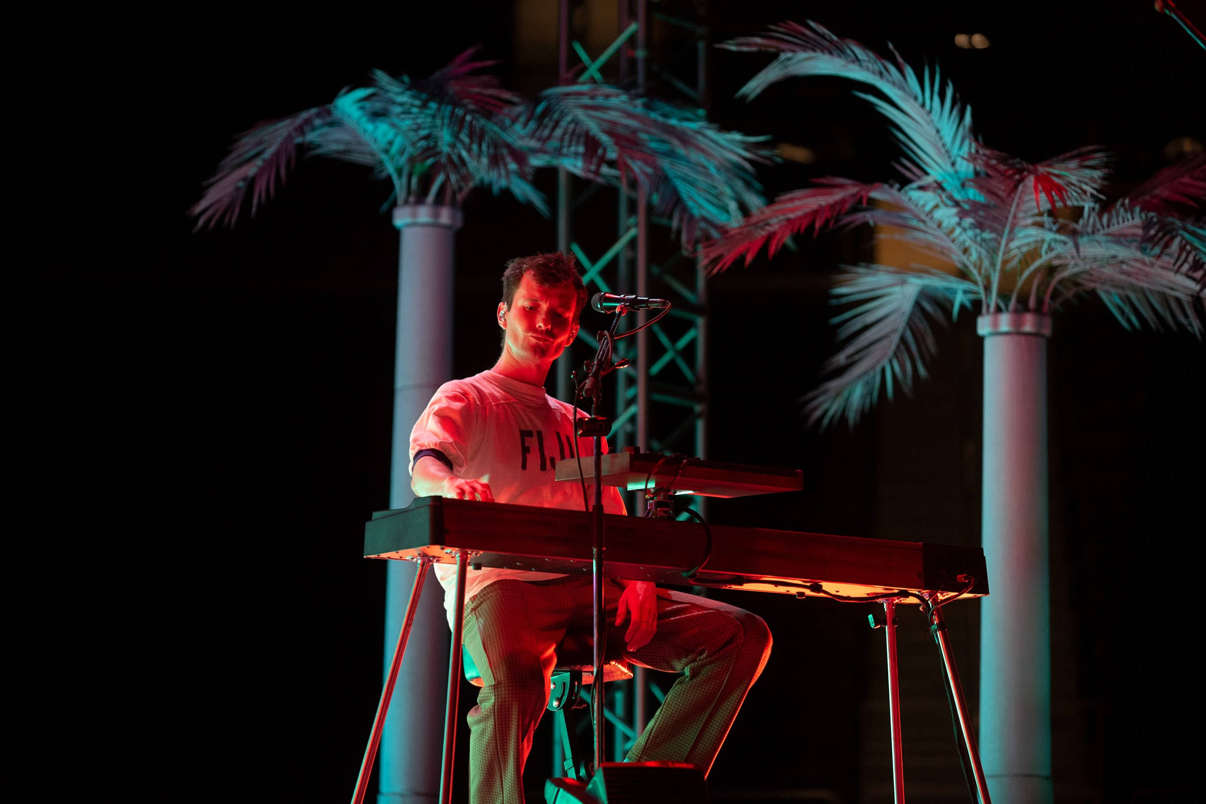 Glass Animals 鍵盤手 Drew MacFarlane 壁紙 — 一位音樂家在舞台上，在色彩繽紛的燈光下彈奏鍵盤，背景有棕櫚樹。