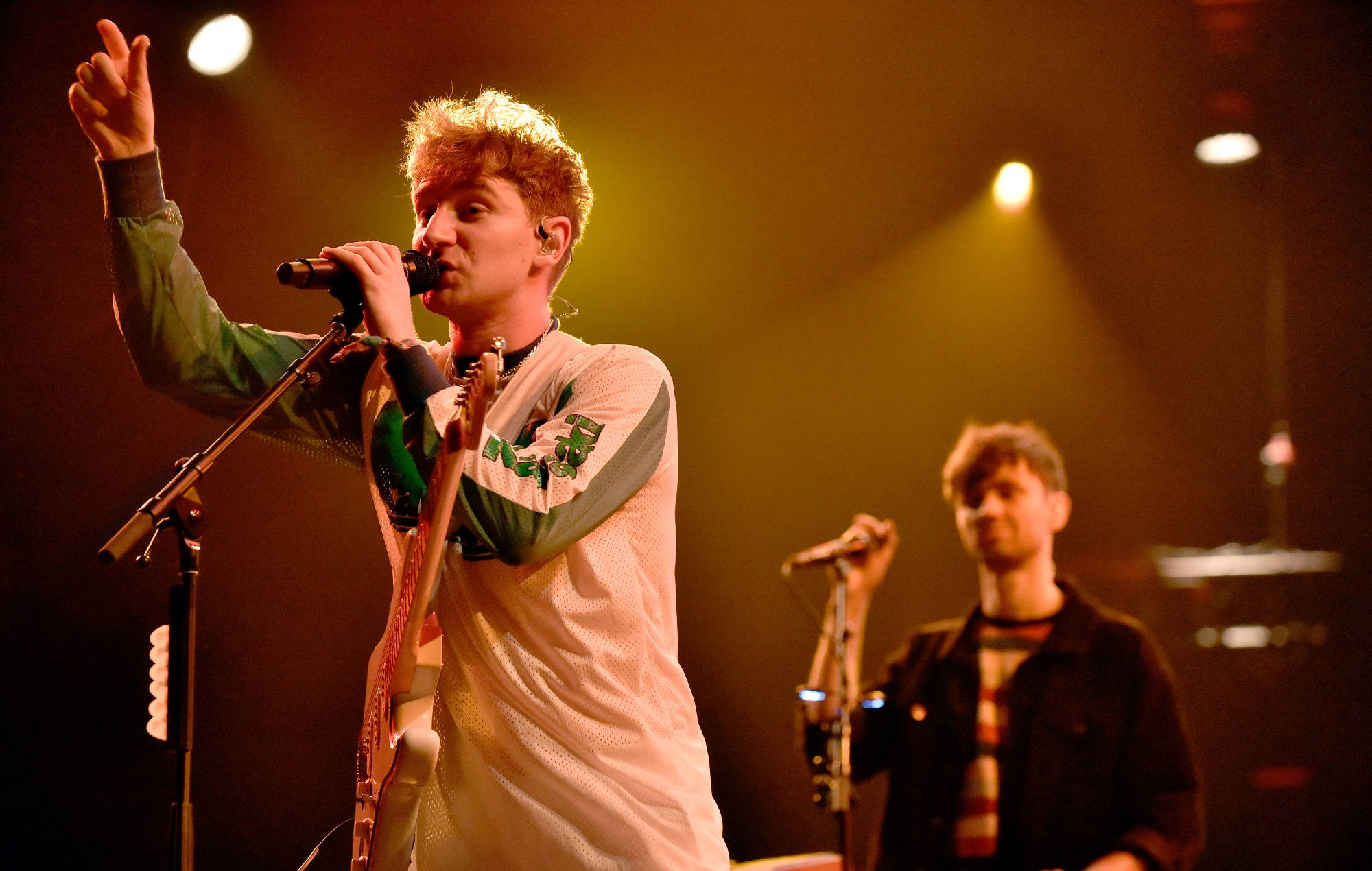 Glass Animals 主唱 Dave Bayley 舞台演唱 壁紙 — 一位金髮男歌手在溫暖的聚光燈下，手持咪高峰和結他，在舞台上充滿激情地表演。