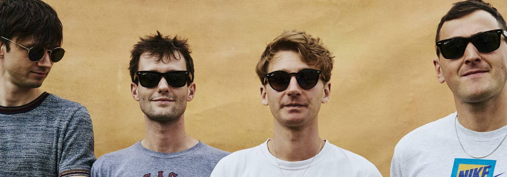 Glass Animals 戴太陽眼鏡 壁紙 — 四個戴太陽眼鏡的男人站在有紋理的芥末黃色牆前，穿著休閒T恤，表情自信。