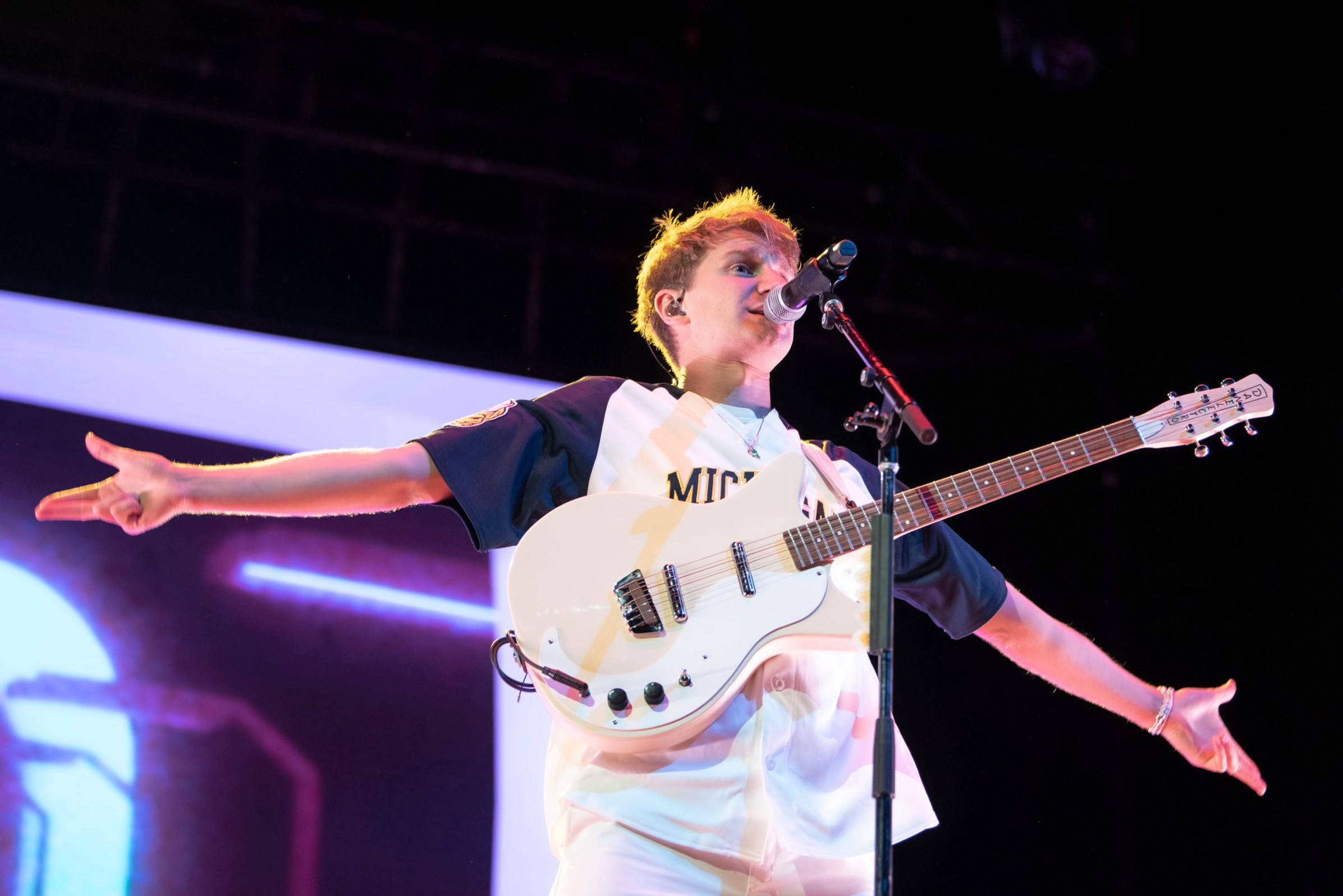 Glass Animals 戴夫 壁紙 — 一位年輕音樂家張開雙臂，在充滿活力的舞台燈光下，熱情地對著咪高峰唱歌，同時手持一把白色電結他。