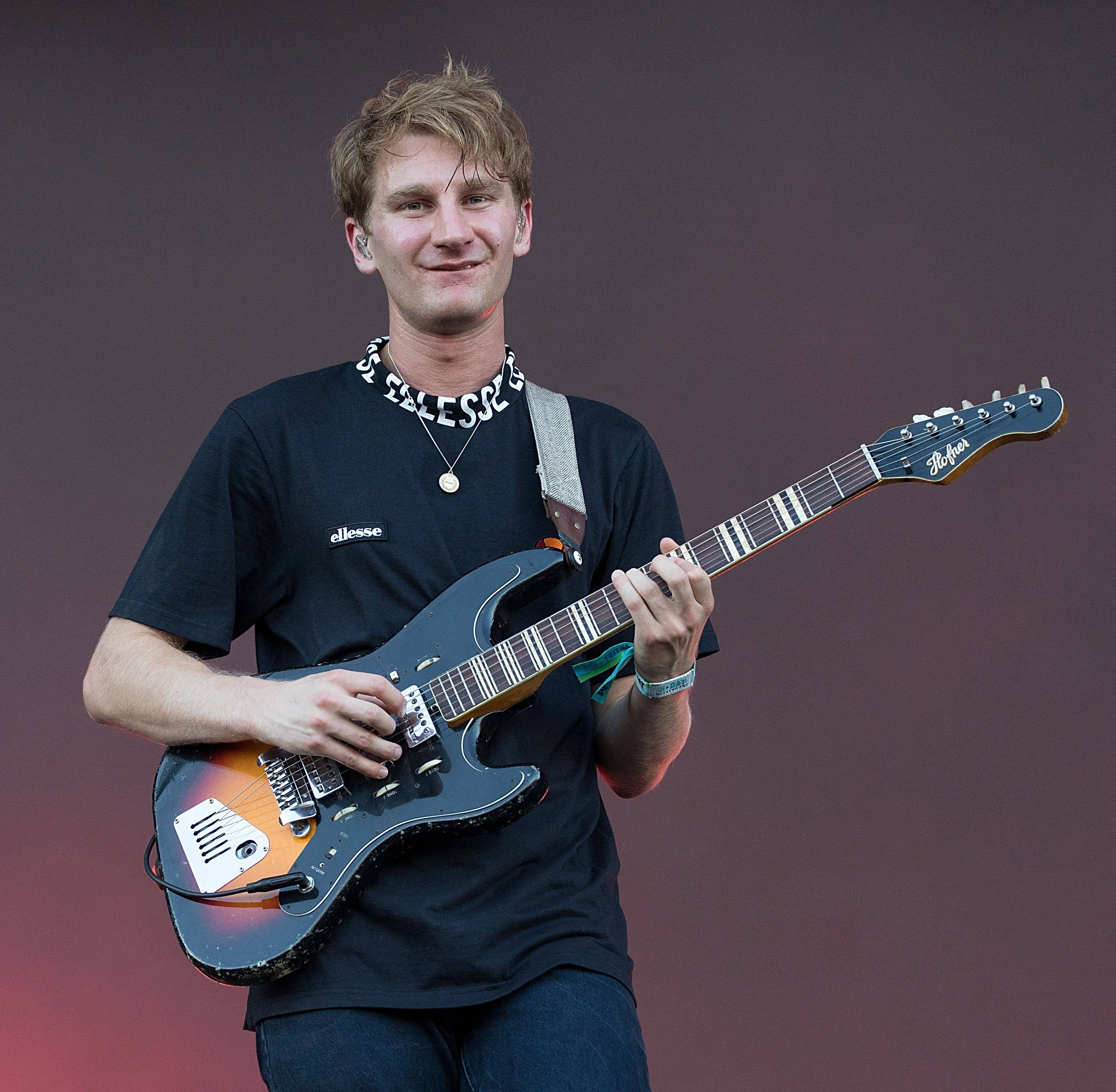 Glass Animals 戴夫·貝利 壁紙 — 一位微笑的年輕男子在紫色和紅色漸變背景的舞台上彈奏一把旭日紋電結他。