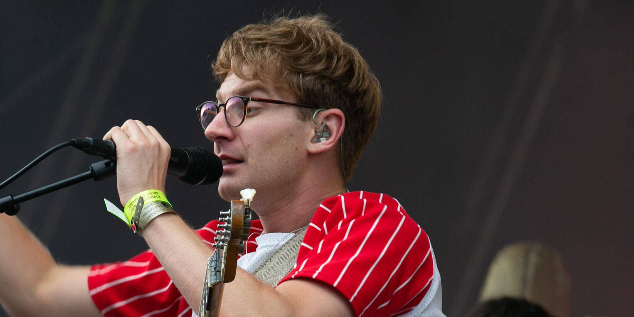 Glass Animals 演唱中 壁紙 — 一位戴眼鏡、身穿紅色條紋襯衫的音樂家在現場音樂會上，一邊拿著結他，一邊對著麥克風唱歌。