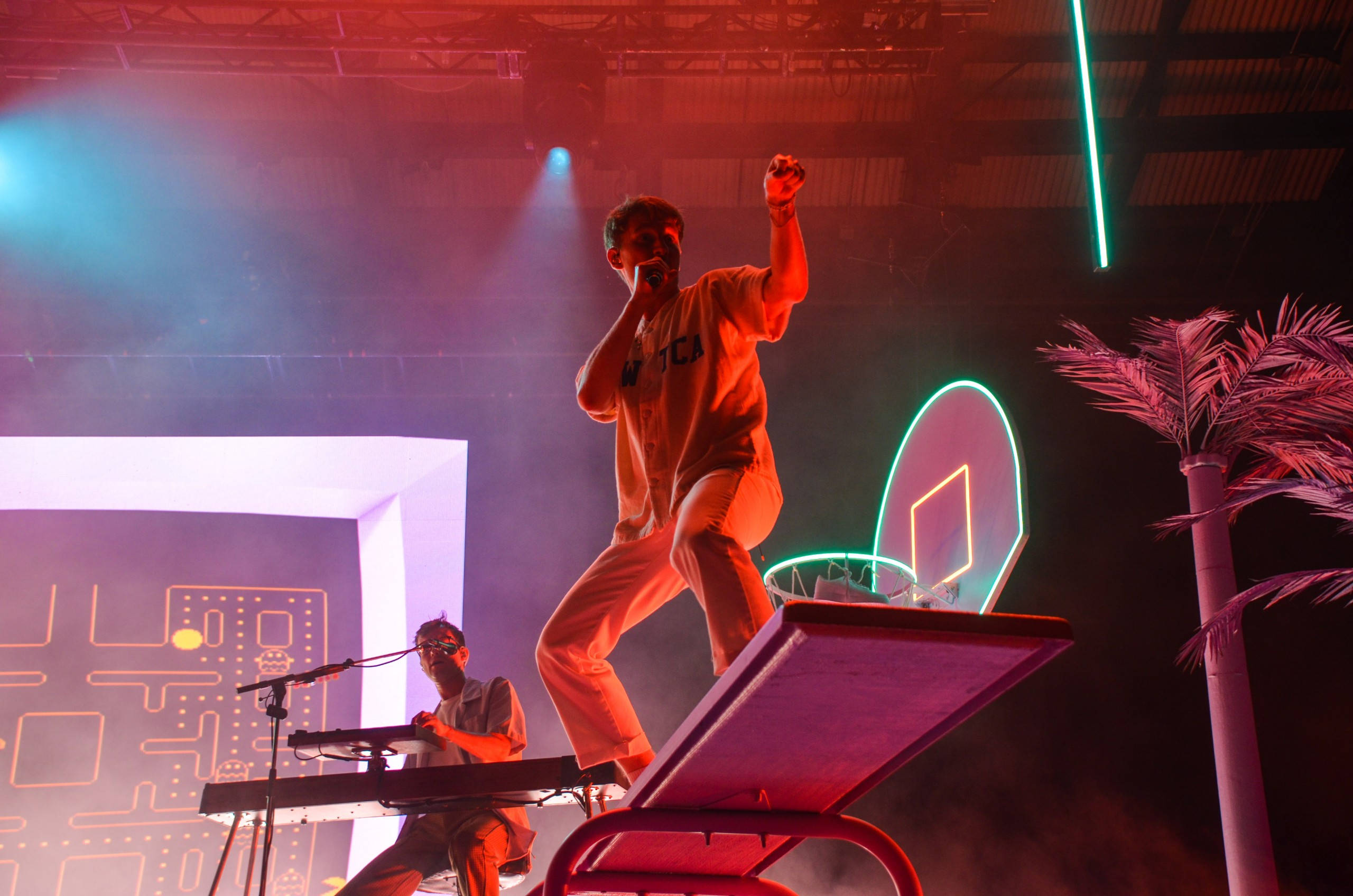 Glass Animals 熱波現場演出 壁紙 — 表演者在霓虹燈下的籃球框舞台上跳躍，而鍵盤手在下方演奏。