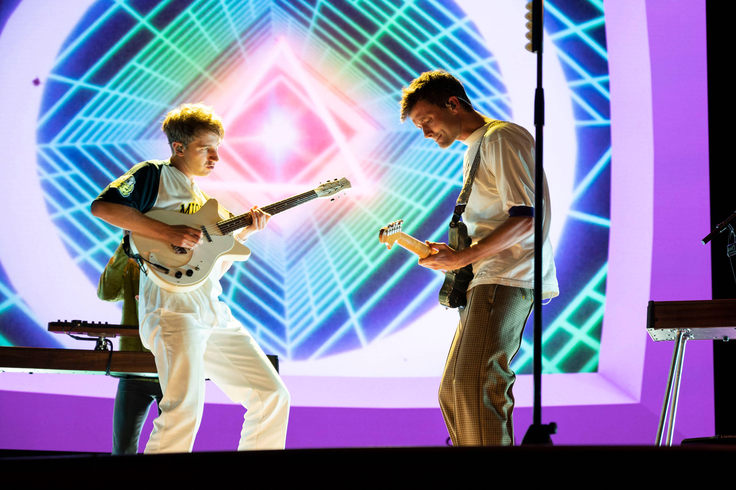 Glass Animals 鍵盤手 Drew MacFarlane 演奏 壁紙 — 兩位音樂家在舞台上，在充滿活力的霓虹幾何燈光下演奏電結他。