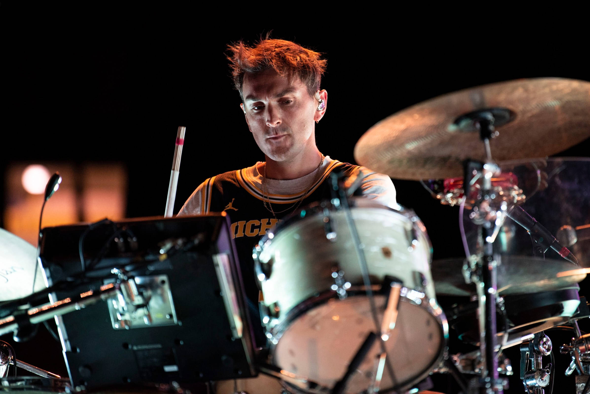 Glass Animals 鼓手 Joe Seaward 壁紙 — 一位專注的鼓手，頭髮凌亂，在黑暗背景的舞台燈光下演奏一套鼓。