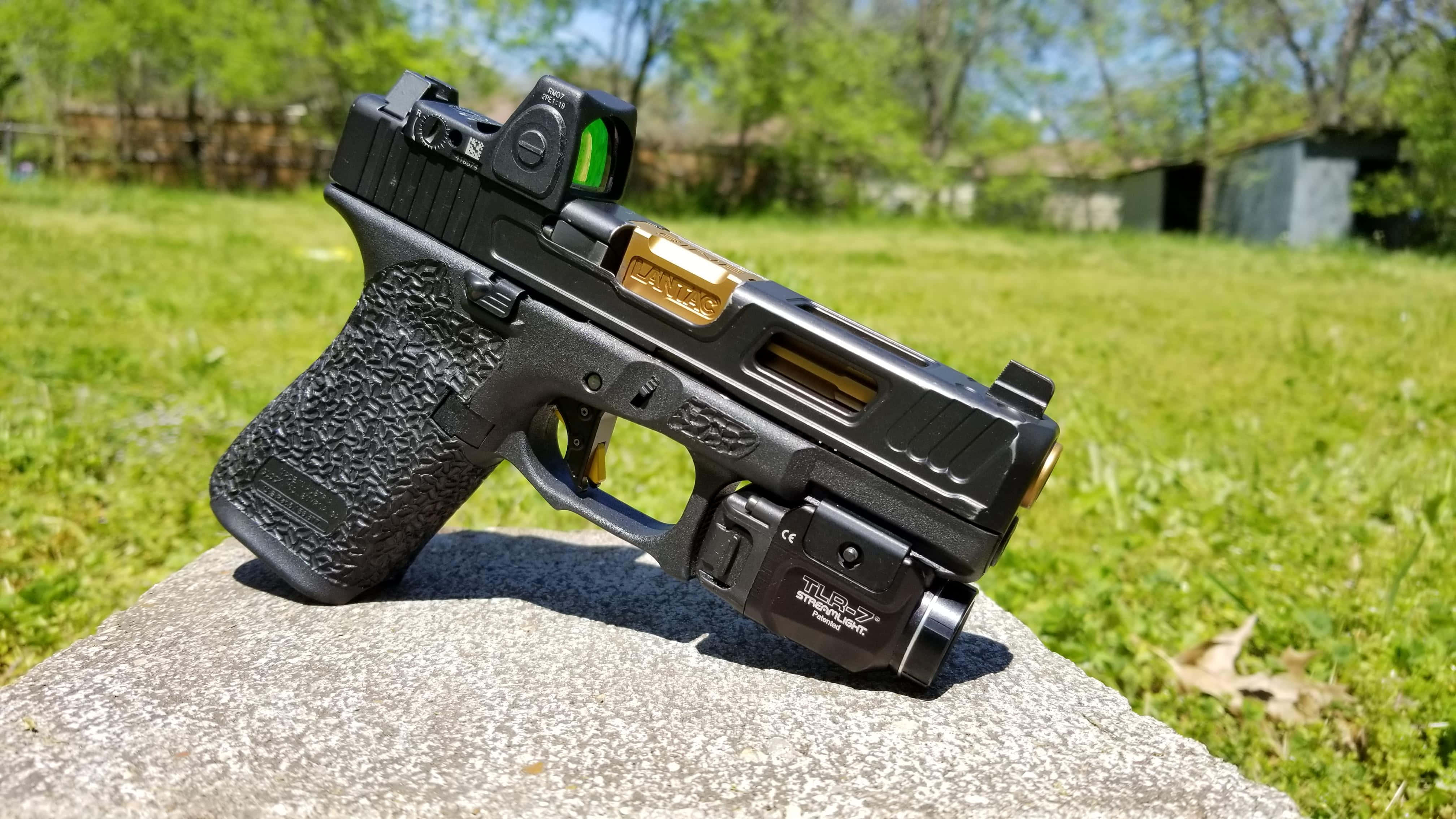 Glock 19 —— 理想的日常攜帶手槍 壁紙 — 一把黑色和金色的手槍配備紅點瞄準器，擱在草地庭院中一塊陽光照耀的石頭上。