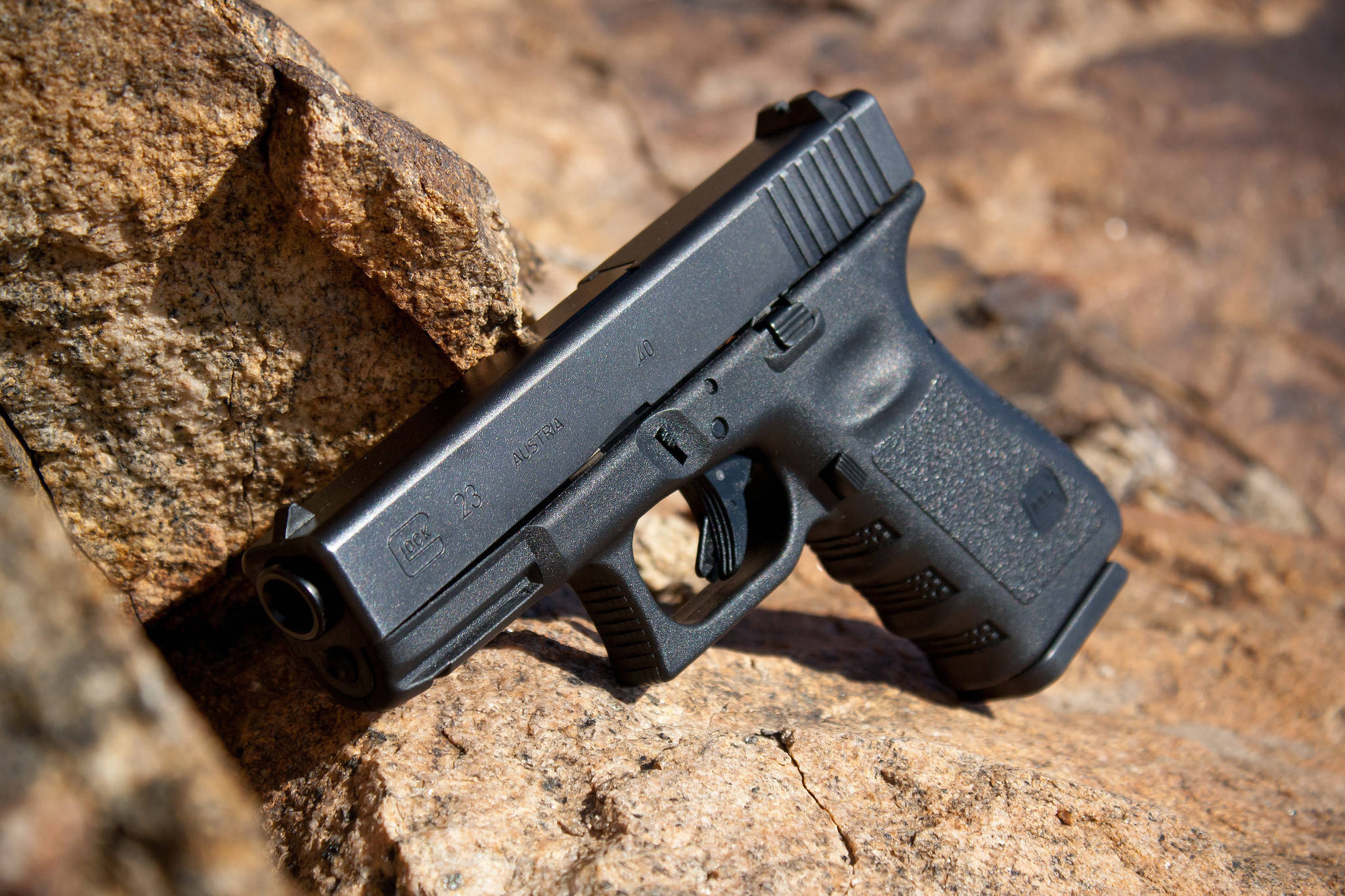 戰術黑色Glock 23 壁紙 — 一把黑色手槍擱在陽光照耀的棕色岩石上，處於崎嶇的戶外環境。
