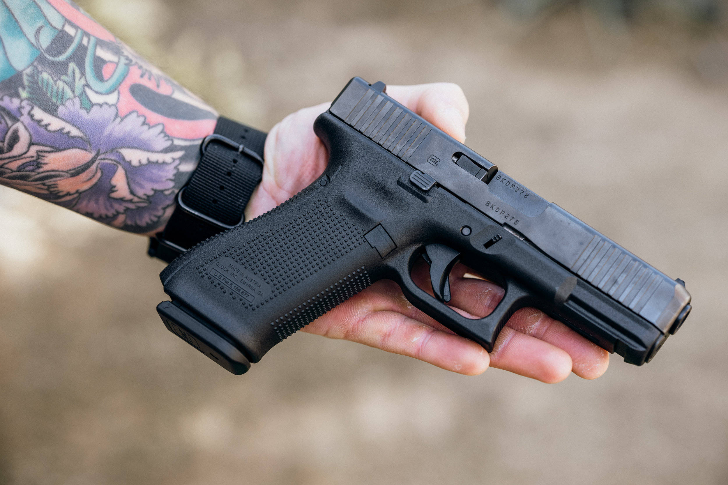 黑色Glock 45手槍 壁紙 — 一隻帶有彩色紋身前臂的手，在模糊的泥土色背景前握著一把黑色半自動手槍。