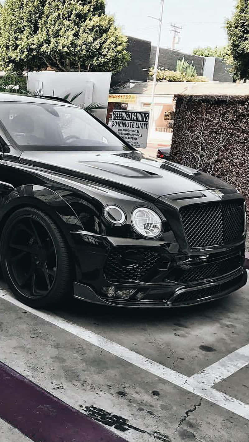Bentley Bentayga 桌布 壁紙 — 一輛亮黑色賓利Bentayga停泊在陽光普照的城市空地，背景有樹木和預留停車標誌。
