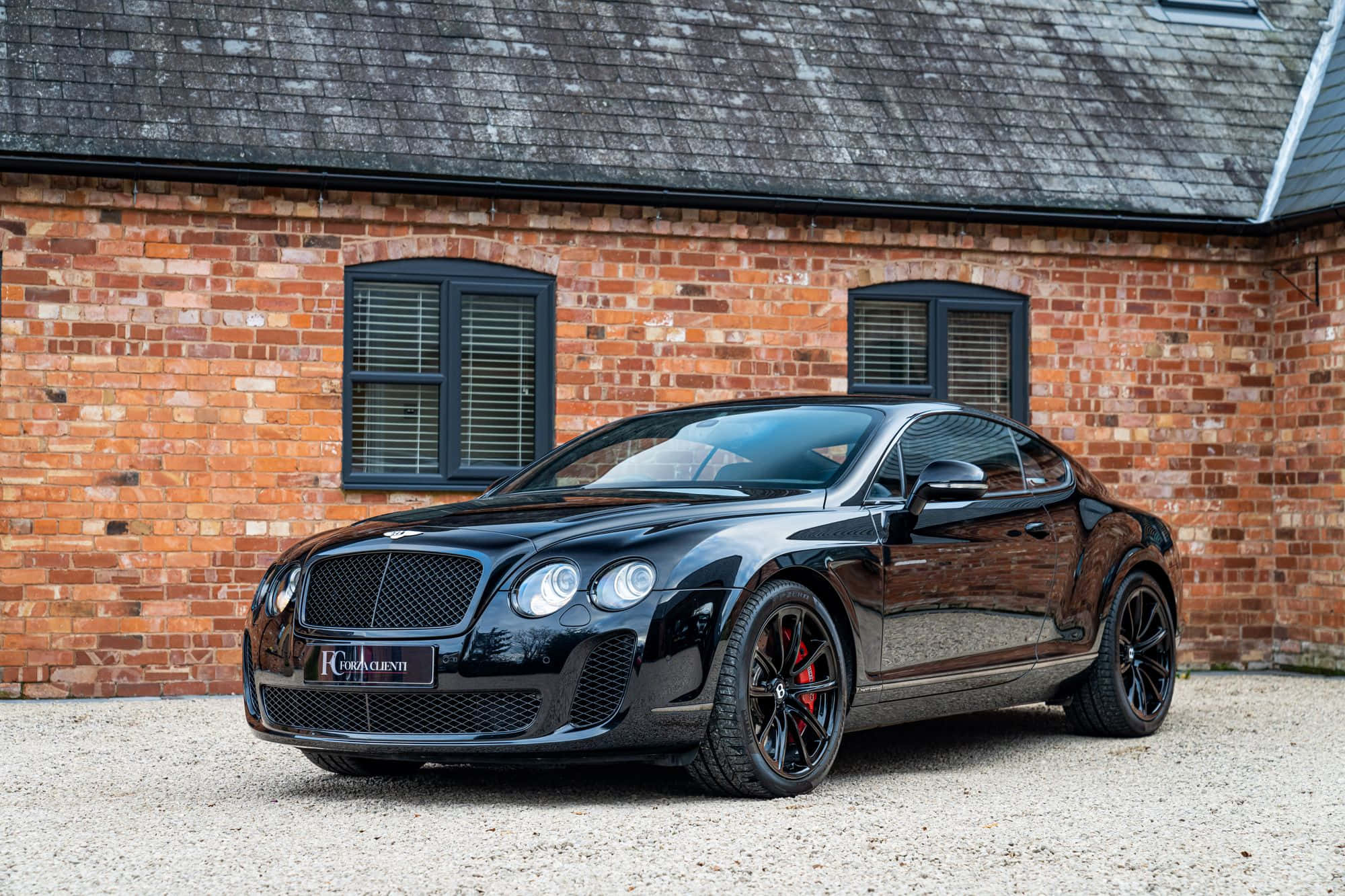 Bentley Continental Supersports 桌布 壁紙 — 一輛亮黑色賓利 Continental GT 雙門跑車停泊在紅磚建築前的碎石地上，建築物有深色窗戶。