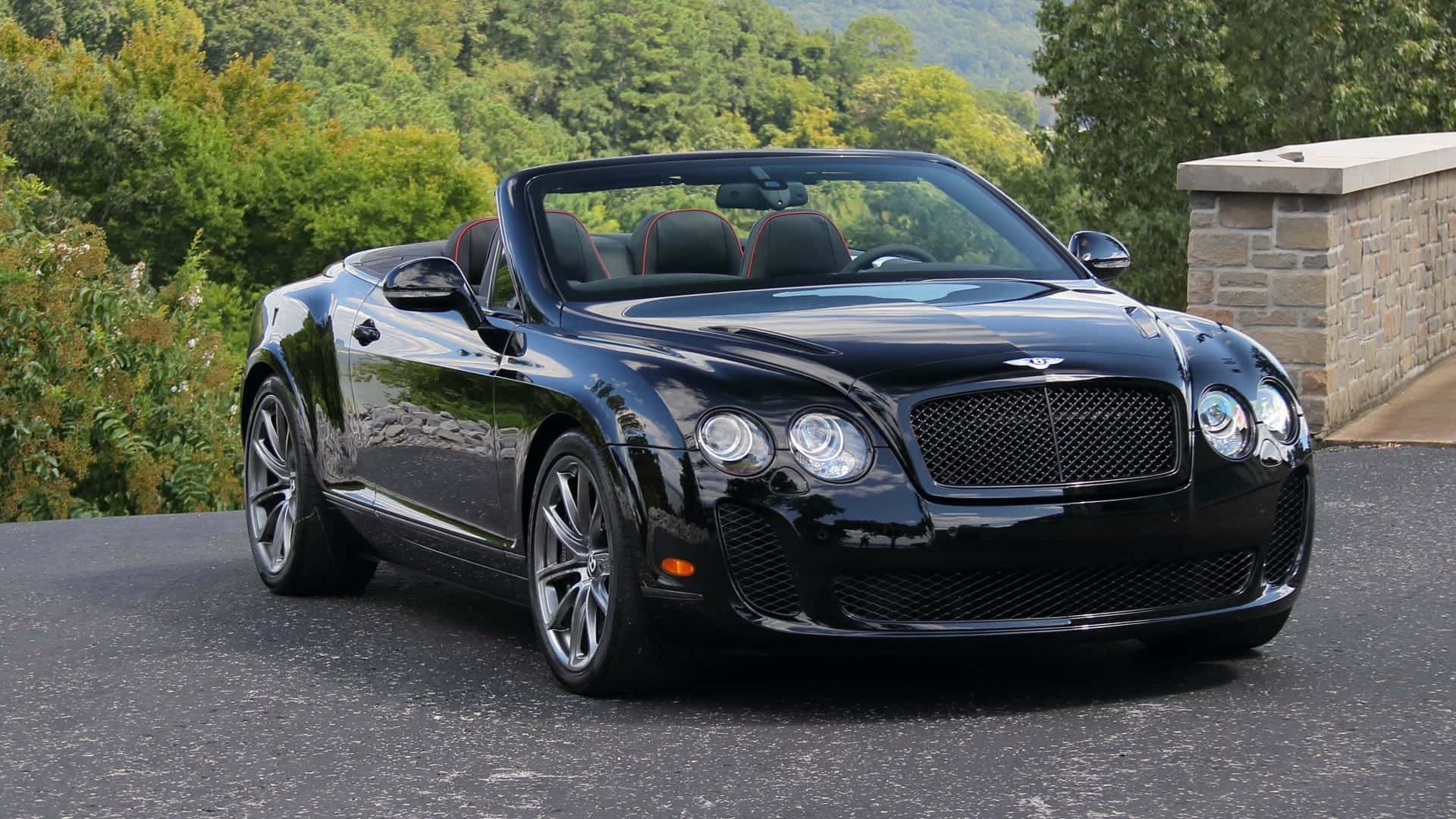 Bentley Continental Supersports 桌布 壁紙 — 一架亮黑色賓利開篷車停泊在瀝青路上，背景有茂密的綠樹和石牆。