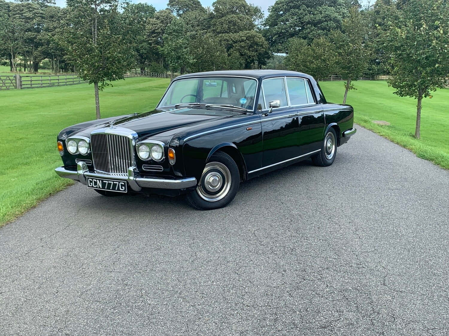 Bentley T1 桌布 壁紙 — 一輛亮黑色賓利S1轎車停泊在碎石車道上，周圍是茂密的綠色草坪和樹木，天空晴朗。